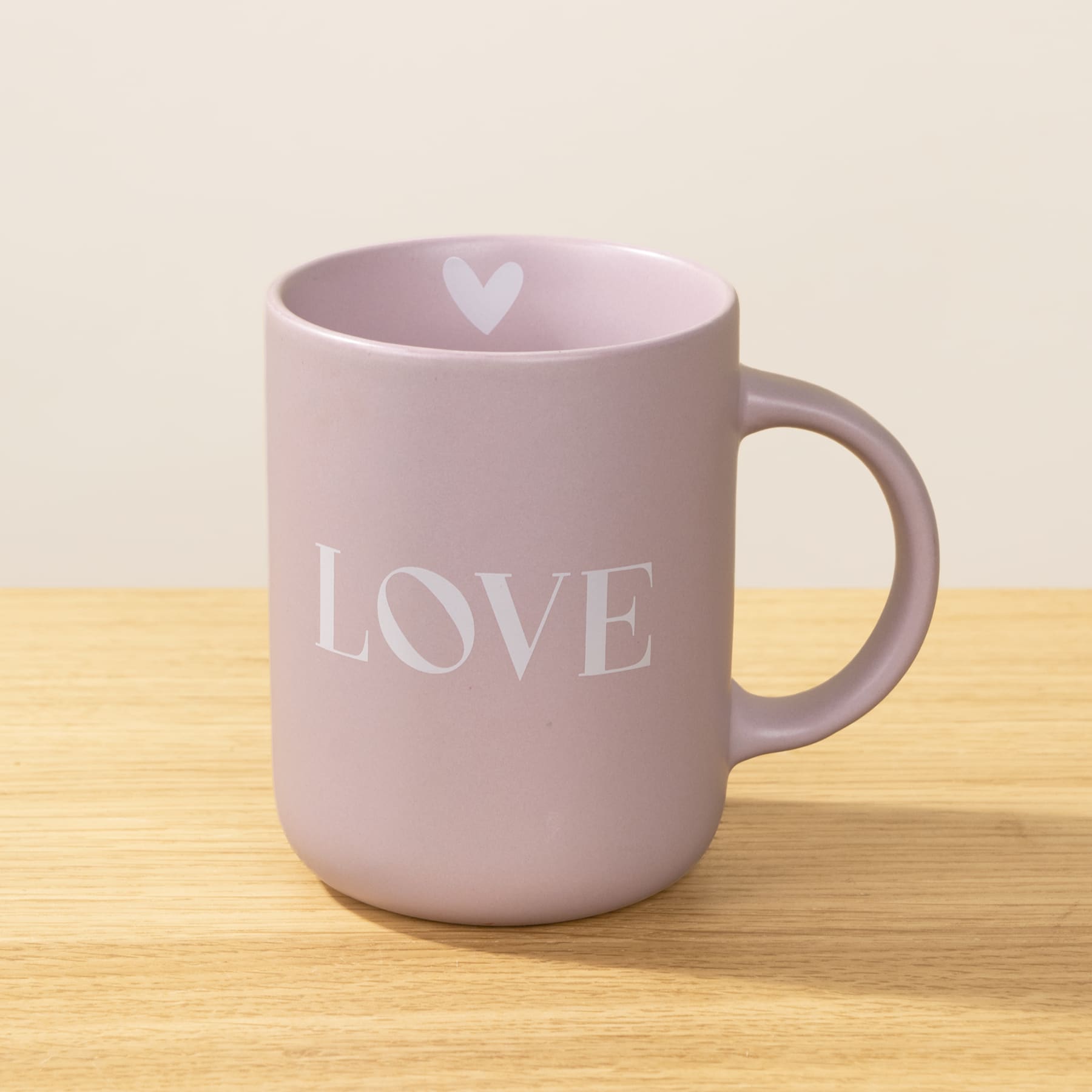 Eine hellrosa Keramiktasse mit dem Aufdruck „LOVE“ steht auf einer Holzoberfläche vor einem schlichten Hintergrund.