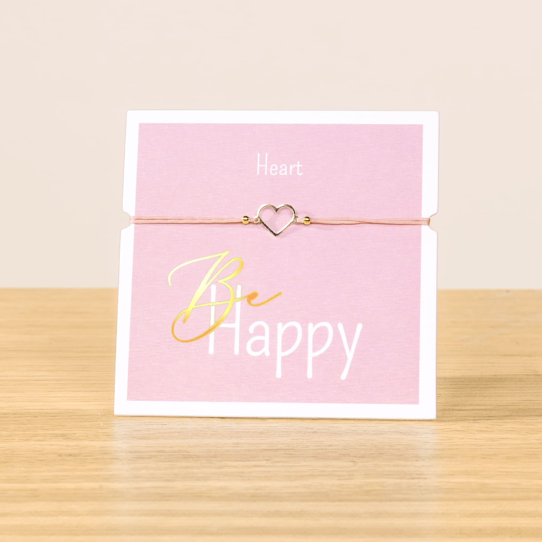 Auf einer Holzoberfläche liegt eine rosa Karte mit dem Text „Be happy“ und einem herzförmigen Anhänger an einer Schnur.