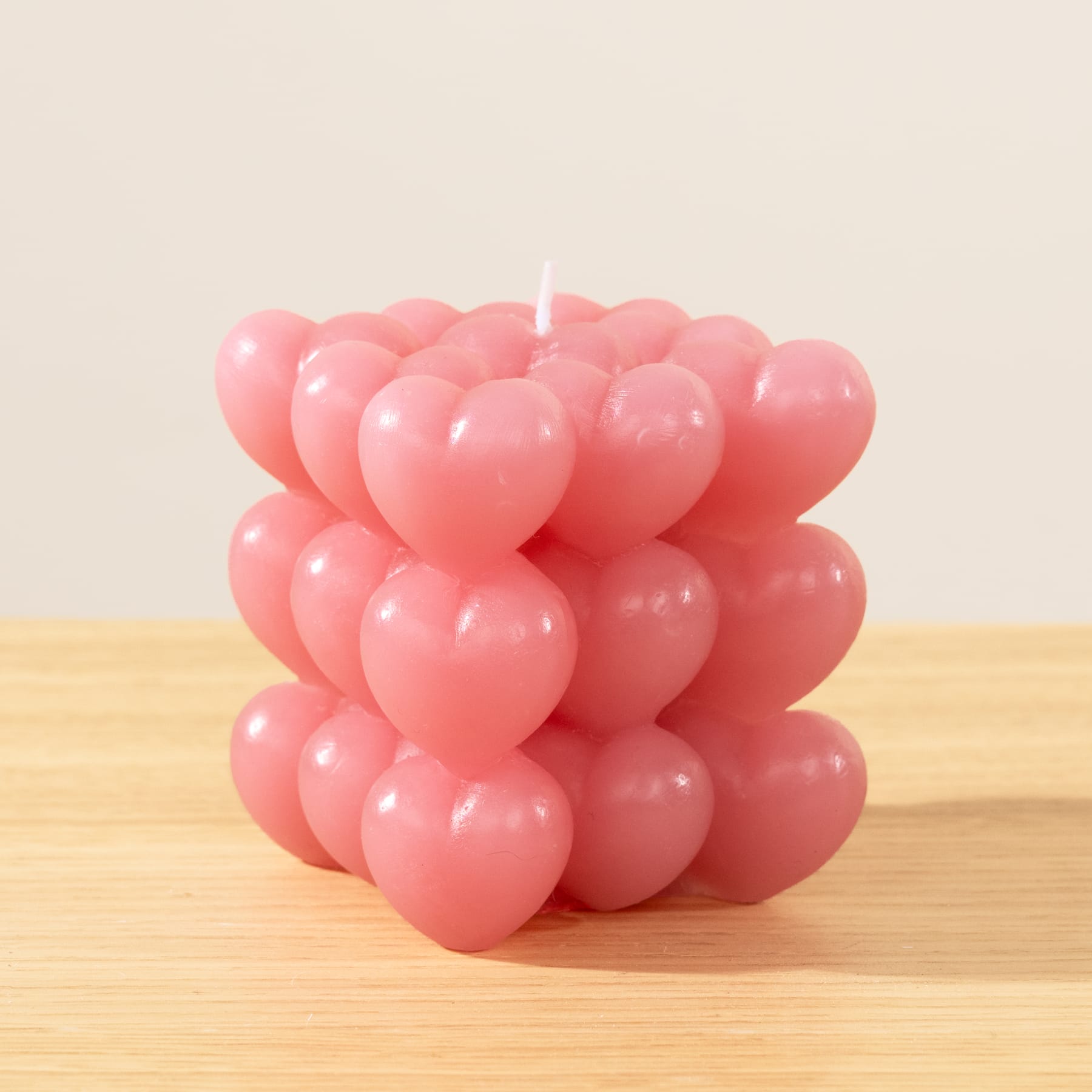Eine würfelförmige Kerze aus rosa herzförmigen Wachsstücken steht auf einer Holzoberfläche vor einem schlichten Hintergrund.