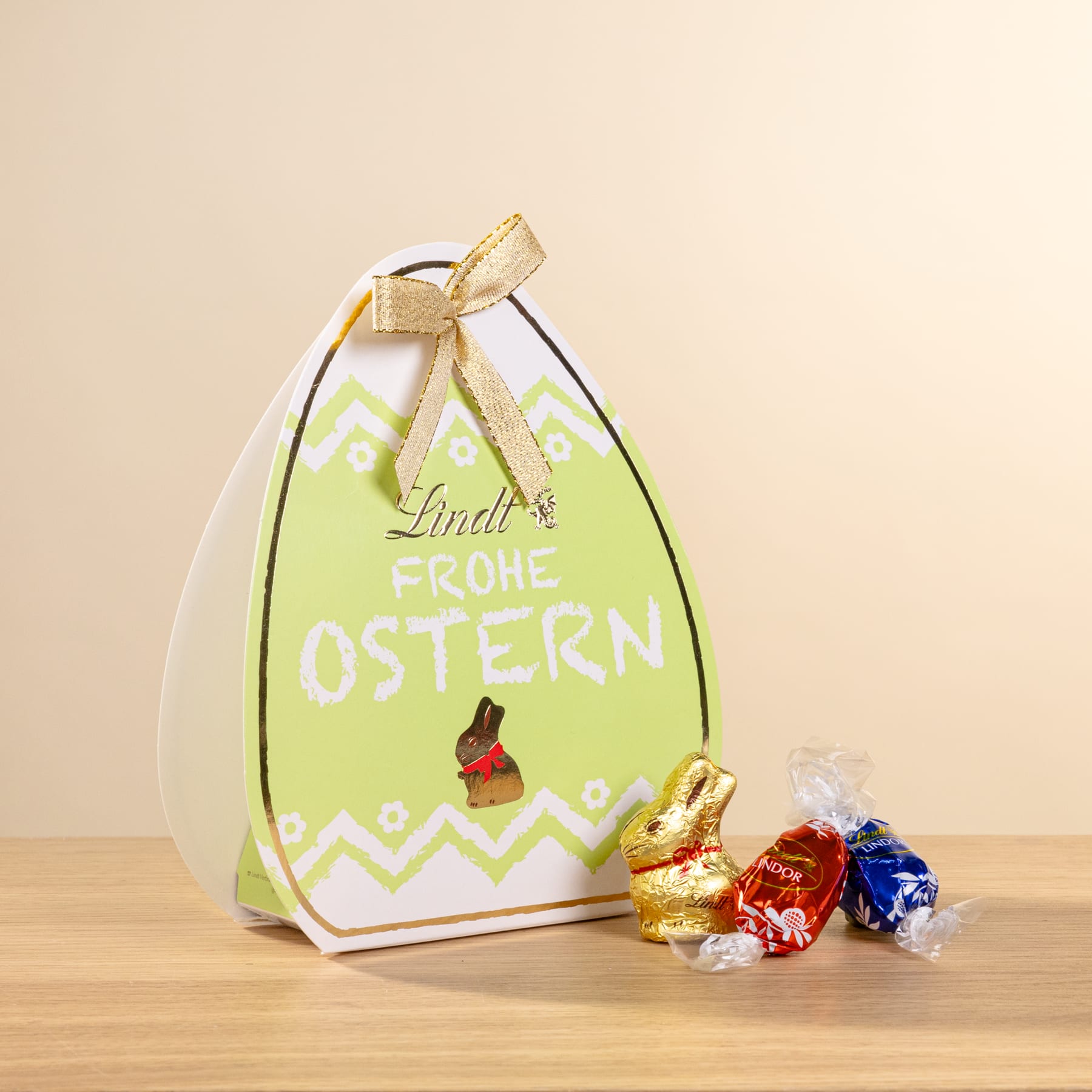 Auf einer Holzfläche steht eine grüne Geschenkbox mit Ostermotiv und goldener Schleife, daneben liegen mehrere kleine Schokoladenbonbons.