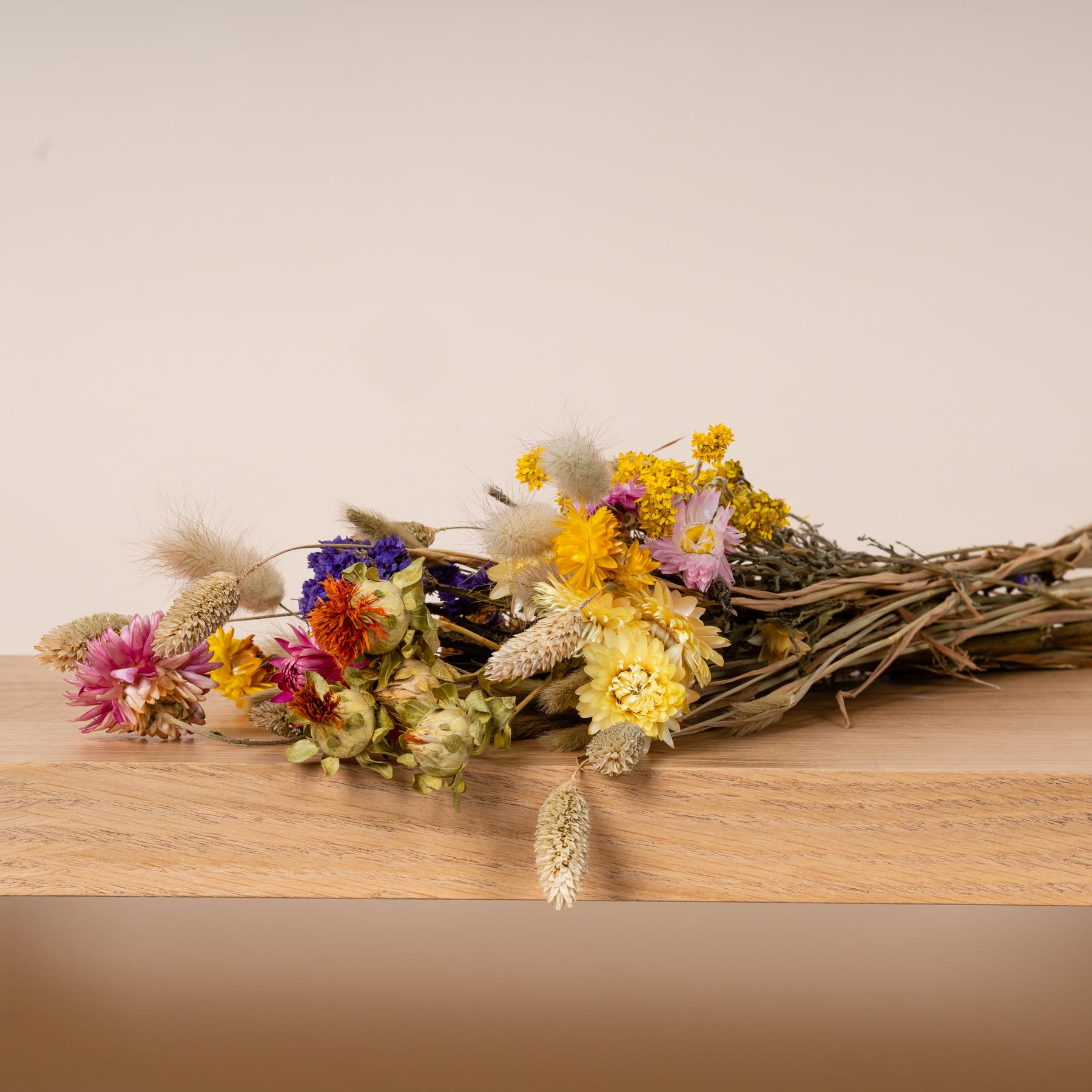 Ein lebendiger Strauß bunter Wildblumen und getrockneter Gräser liegt auf einer Holzoberfläche vor einem neutralen Hintergrund.