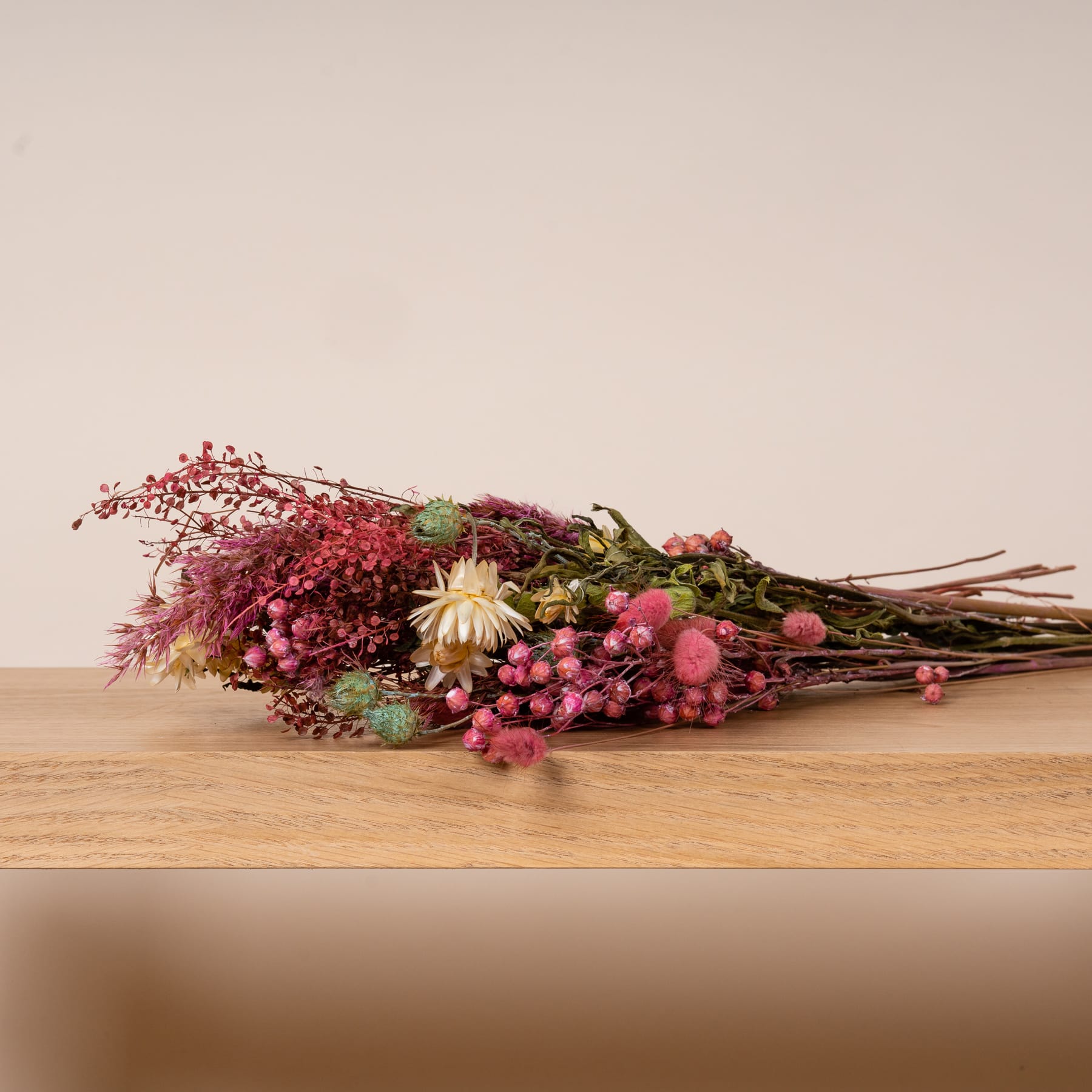 Ein farbenfroher Strauß verschiedener Trockenblumen, darunter rote Beeren, rosa Blüten und ein weißes Gänseblümchen, liegt auf einer Holzoberfläche vor einem neutralen Hintergrund.