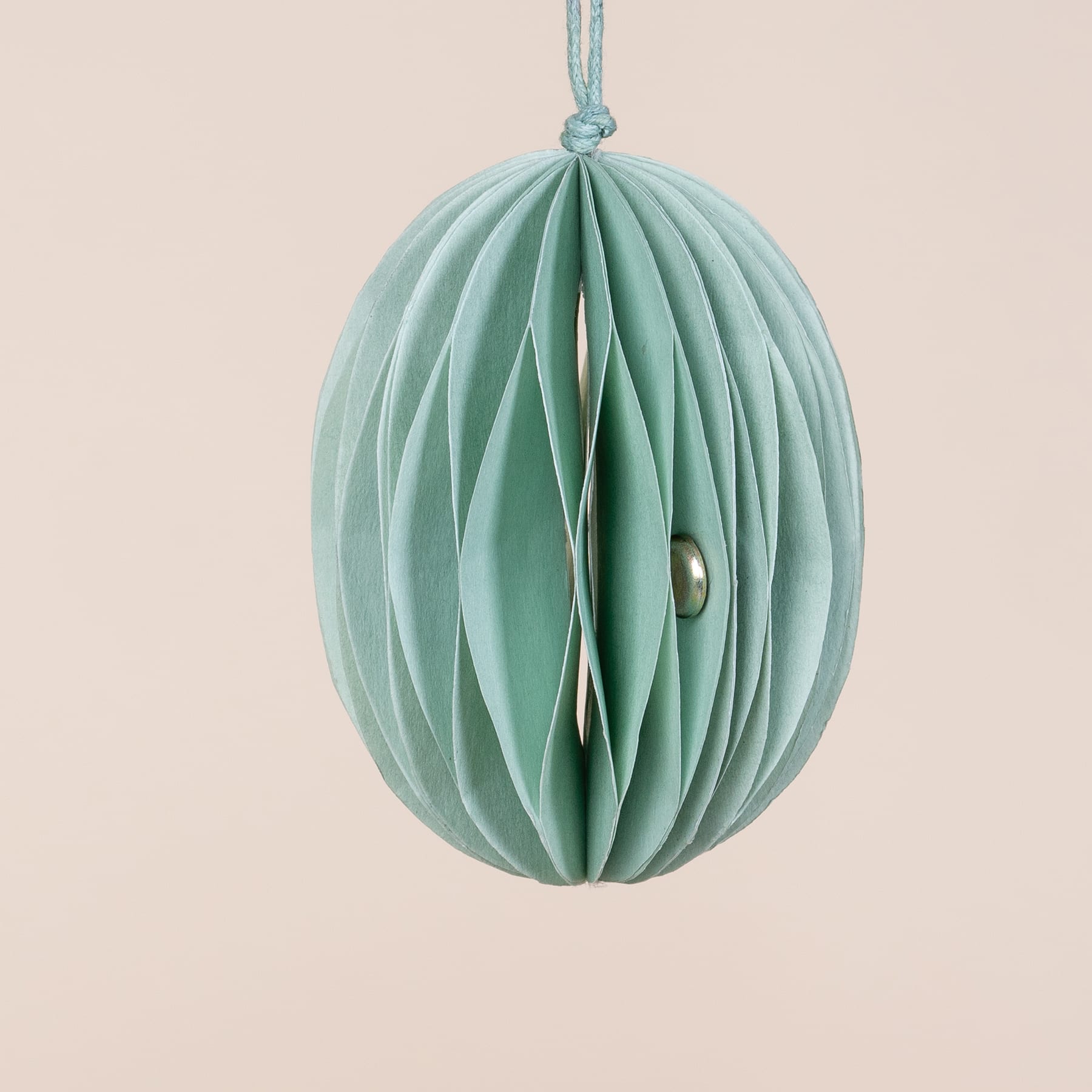 Ein mintgrünes, aus Papier gefaltetes Ornament mit einer Metallperle, die an einer hellblauen Schnur vor einem hellbeigen Hintergrund hängt.