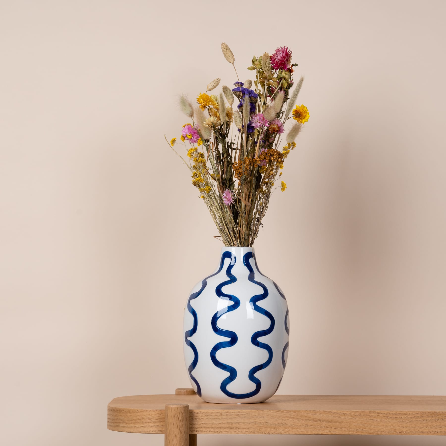 Eine Vase mit wellenförmigem blau-weißem Muster enthält einen farbenfrohen Strauß getrockneter Blumen vor einem neutralen beigen Hintergrund.
