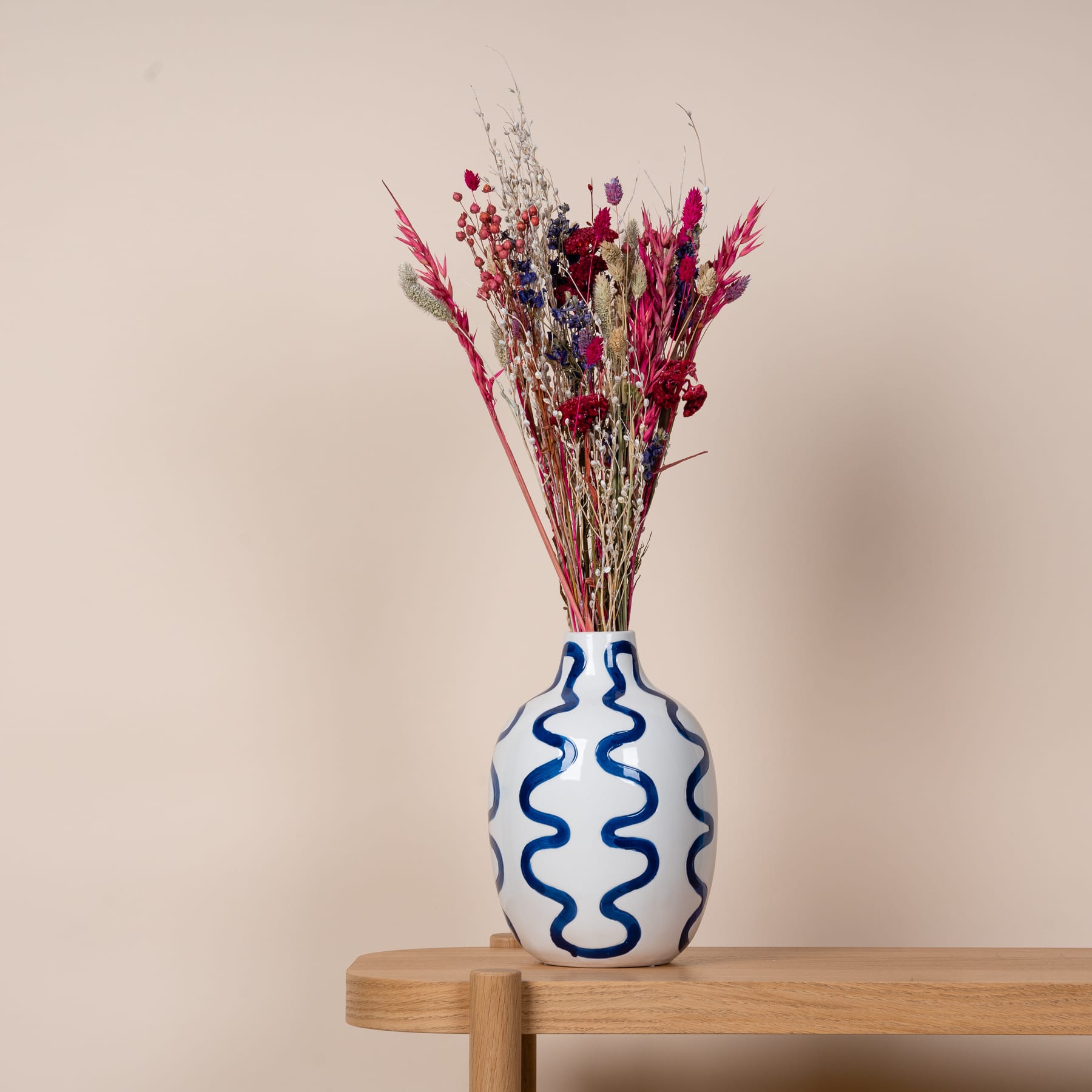 Auf einem Holzregal steht eine Keramikvase mit einem wellenförmigen blau-weißen Muster und enthält einen farbenfrohen Strauß getrockneter roter und violetter Blumen.