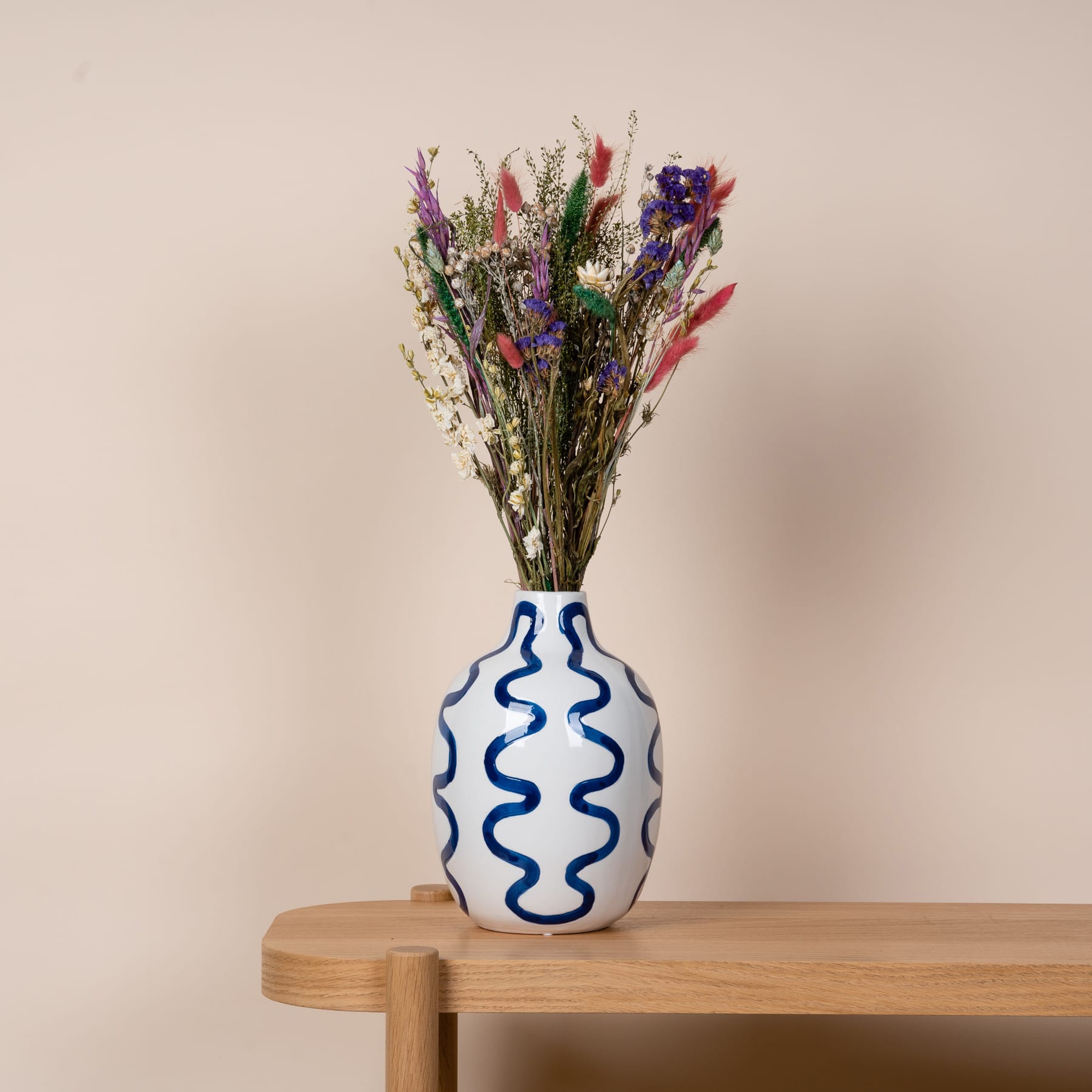 Eine dekorative Keramikvase mit einem wellenförmigen blauen Muster enthält einen lebendigen Strauß bunter Wildblumen vor einem neutralen beigen Hintergrund.