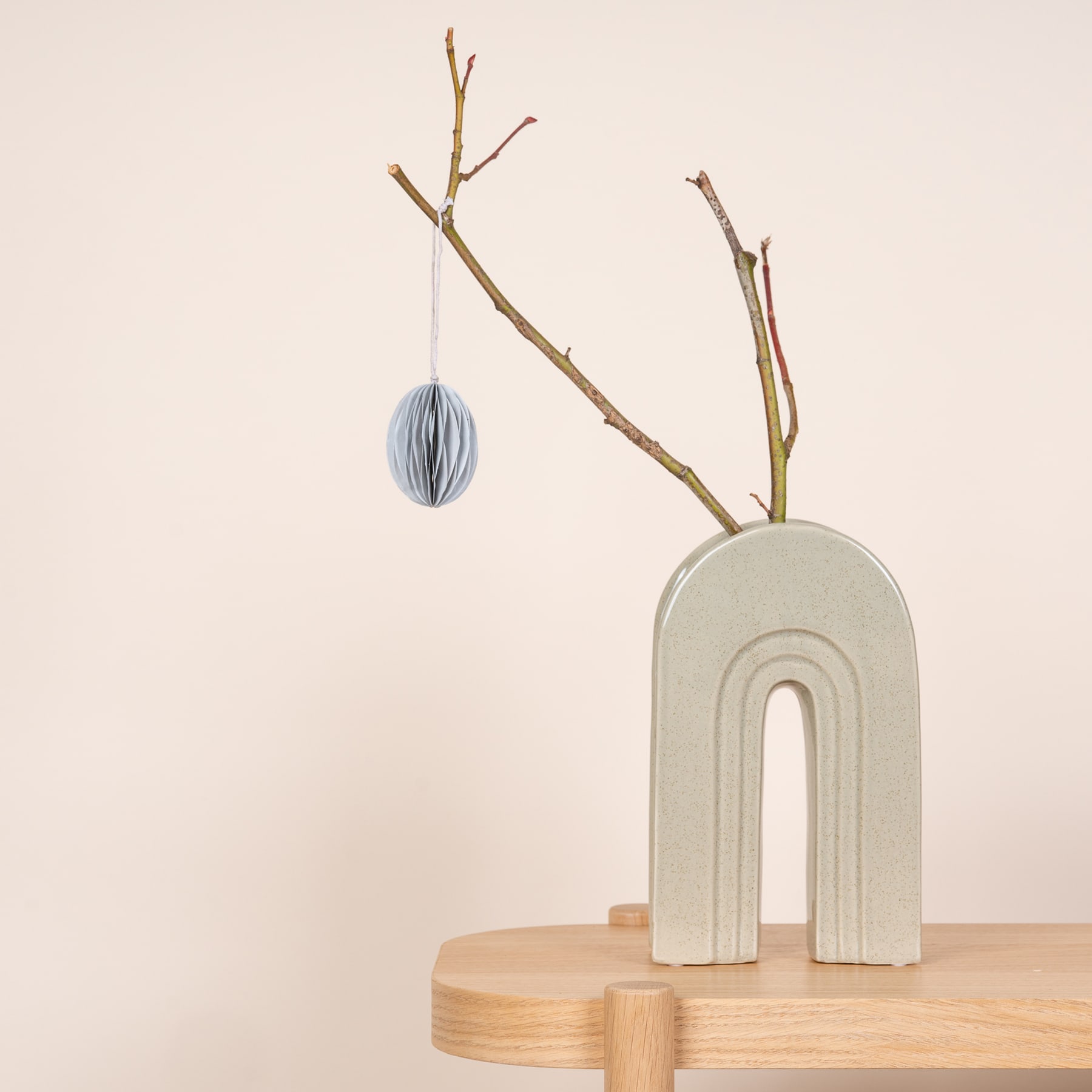 Ein minimalistisches Arrangement mit einer weißen Keramikvase mit hängendem Papierornament, platziert auf einem Holzregal vor einem neutralen Hintergrund.