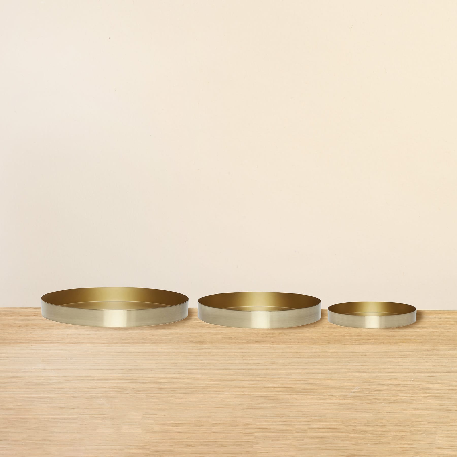 Drei runde, goldfarbene Tabletts oder Teller sind auf einer Holzoberfläche vor einem schlichten, hellen Hintergrund angeordnet.