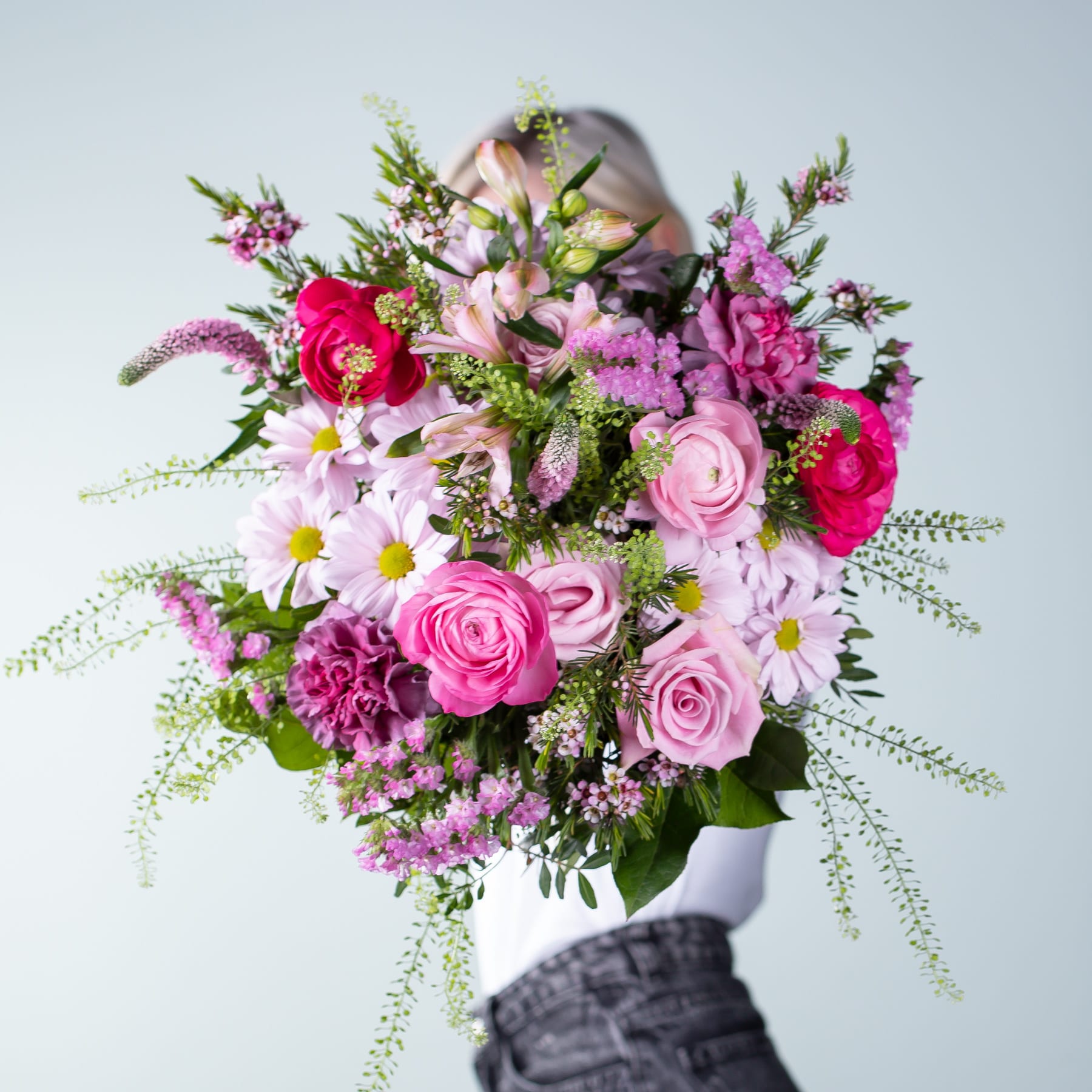 Ein farbenfroher Strauß verschiedener rosa, roter und weißer Blumen, darunter Rosen, Nelken und Gänseblümchen, wird vor einem schlichten grauen Hintergrund gehalten.