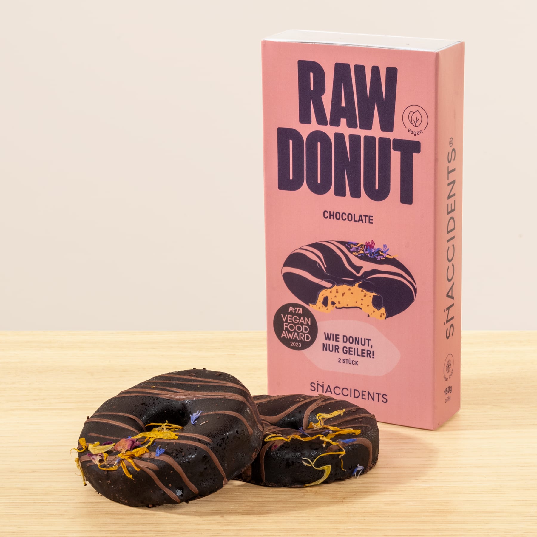 Das Bild zeigt eine rosafarbene Schachtel mit der Aufschrift „RAW DONUT“ und Schokoladendonuts auf einer Holzoberfläche im Vordergrund.