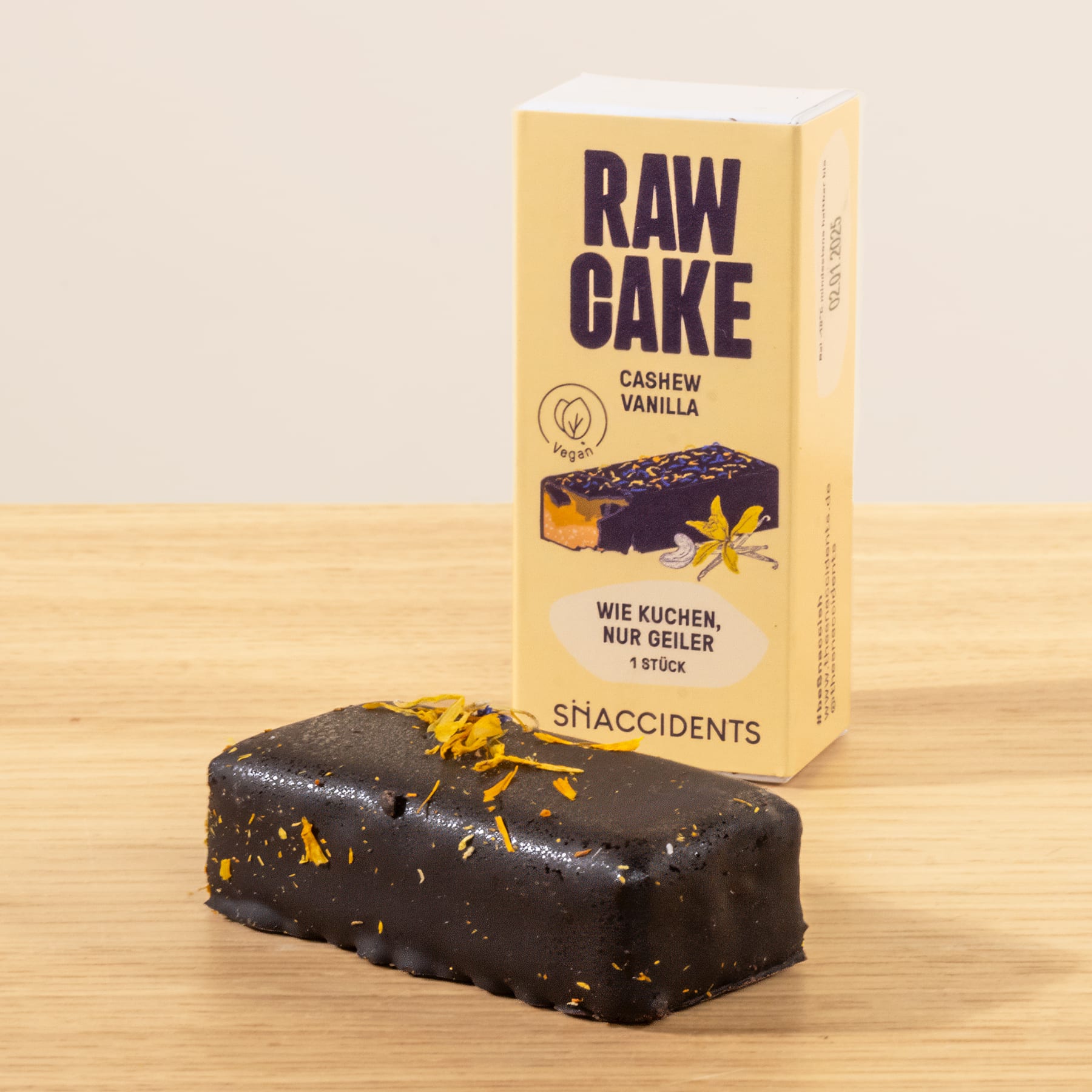 Auf einer Holzfläche steht ein rechteckiger Rohkuchen mit Zartbitterschokoladenüberzug, im Hintergrund eine Schachtel mit der Aufschrift „Raw Cake Cashew Vanilla“.