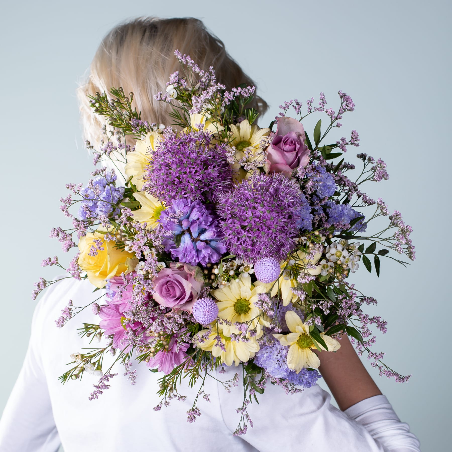 Ein leuchtender Strauß bunter Blumen, darunter violette Chrysanthemen, gelbe Narzissen und rosa Rosen, gehalten von einer Person in einem weißen Hemd vor einem schlichten Hintergrund.