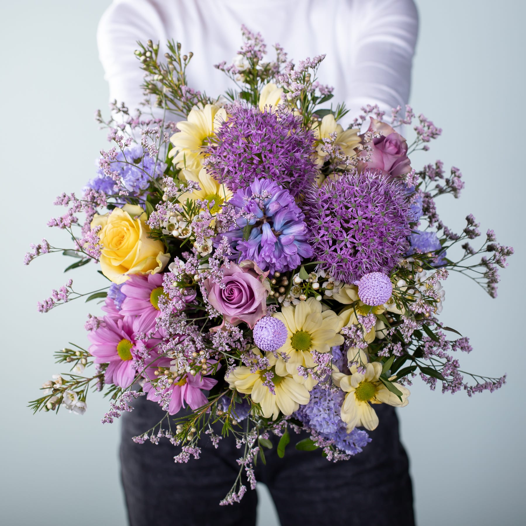 Ein leuchtender Strauß bunter Blumen, darunter lila Allium, gelbe Narzissen und rosa Rosen, wird vor einem schlichten Hintergrund in den Händen einer Person gehalten.