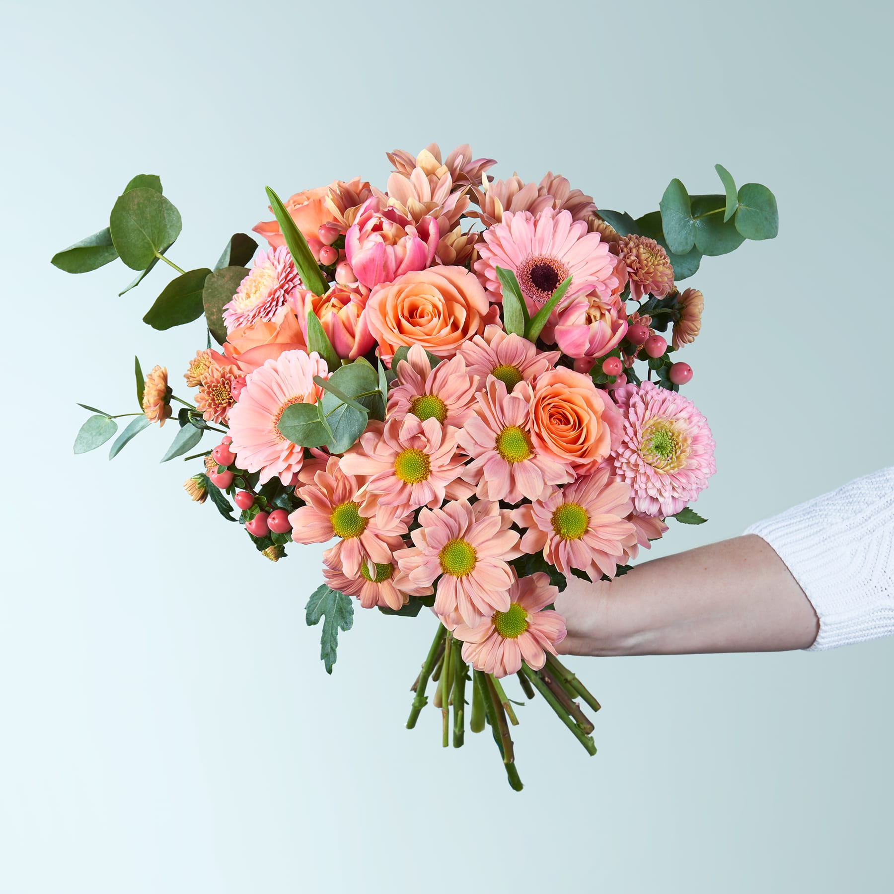 Ein leuchtender Strauß rosa und orangefarbener Blumen, darunter Rosen und Gänseblümchen, wird vor einem hellblauen Hintergrund in den Händen einer Person gehalten.
