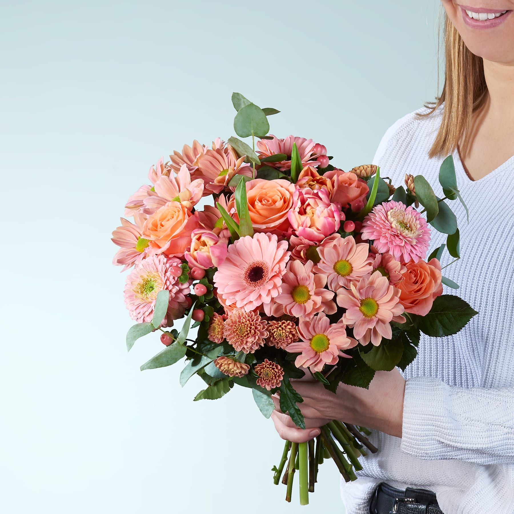 Ein farbenfroher Strauß rosa und orangefarbener Blumen, darunter Rosen, Gerbera und Chrysanthemen, wird von einer Person gehalten, die ein weißes Hemd vor einem schlichten hellblauen Hintergrund trägt.