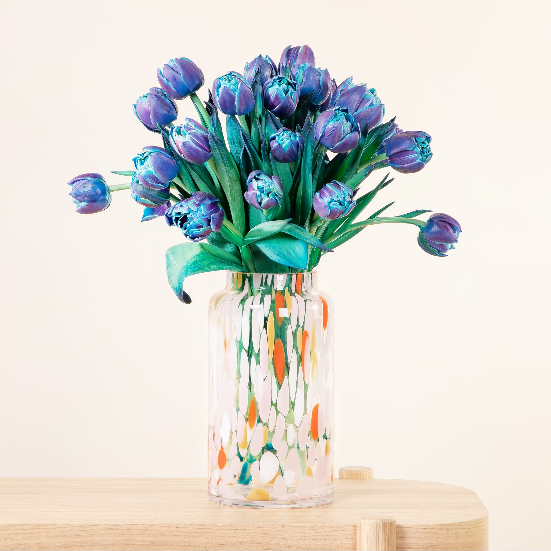 Ein leuchtender Strauß violetter und blauer Tulpen steht in einer bunten, gemusterten Glasvase vor einem schlichten, hellen Hintergrund.