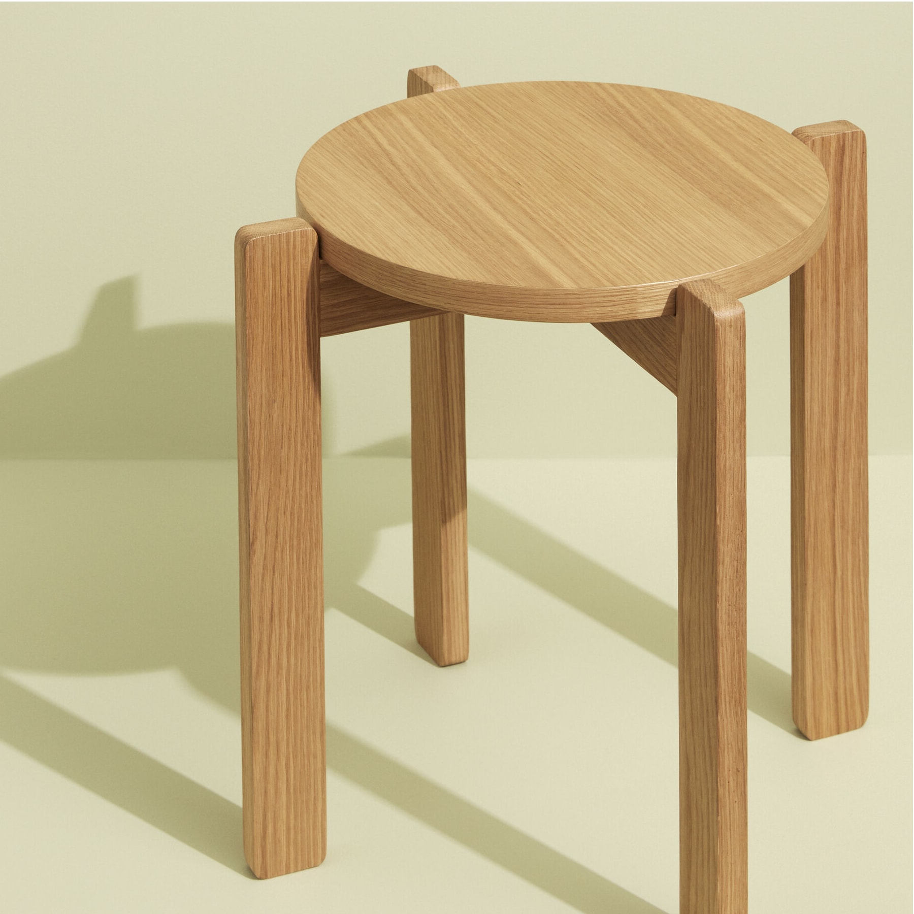 Ein einfacher, runder Holzhocker oder Beistelltisch steht auf einem schlichten, hellen Hintergrund und wirft einen deutlichen Schatten auf die Oberfläche dahinter.
