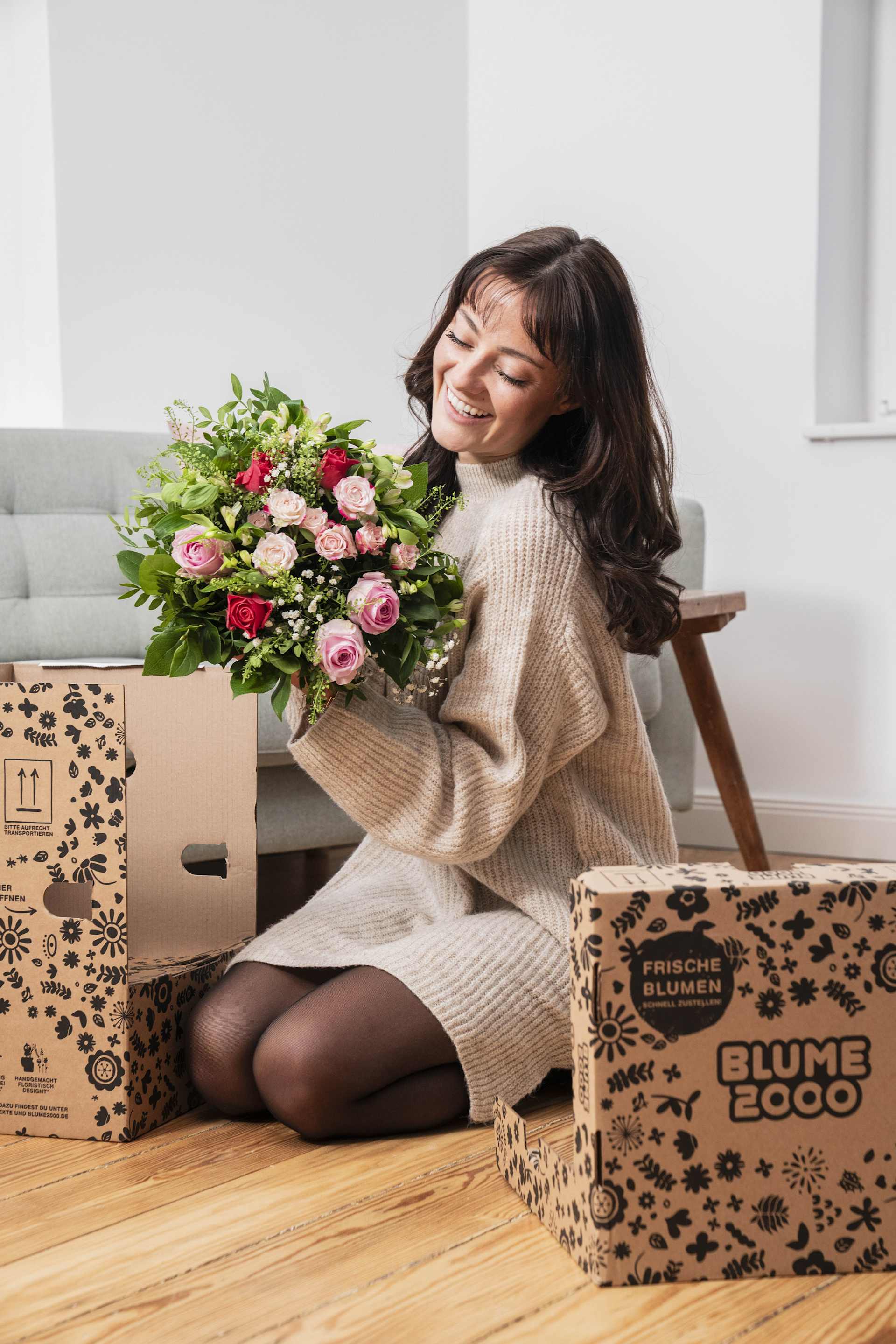 Eine junge Frau in einem kuscheligen Pullover sitzt umgeben von Pappkartons auf dem Boden, hält einen farbenfrohen Blumenstrauß in der Hand und lächelt zufrieden.