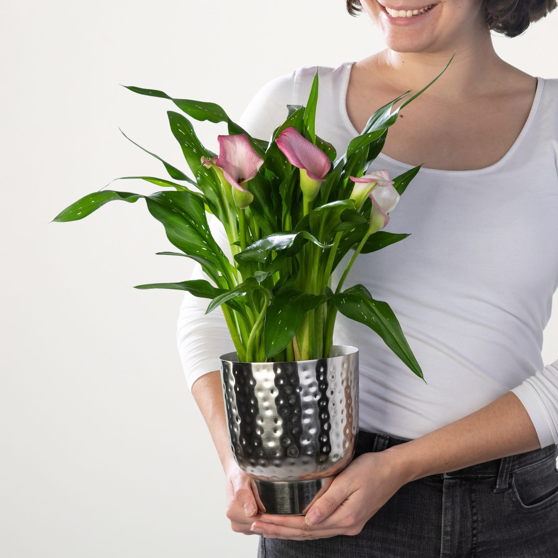 Eine Frau in einem weißen Oberteil hält vor einem schlichten weißen Hintergrund einen Strauß rosa Blumen in einer gepunkteten Vase.