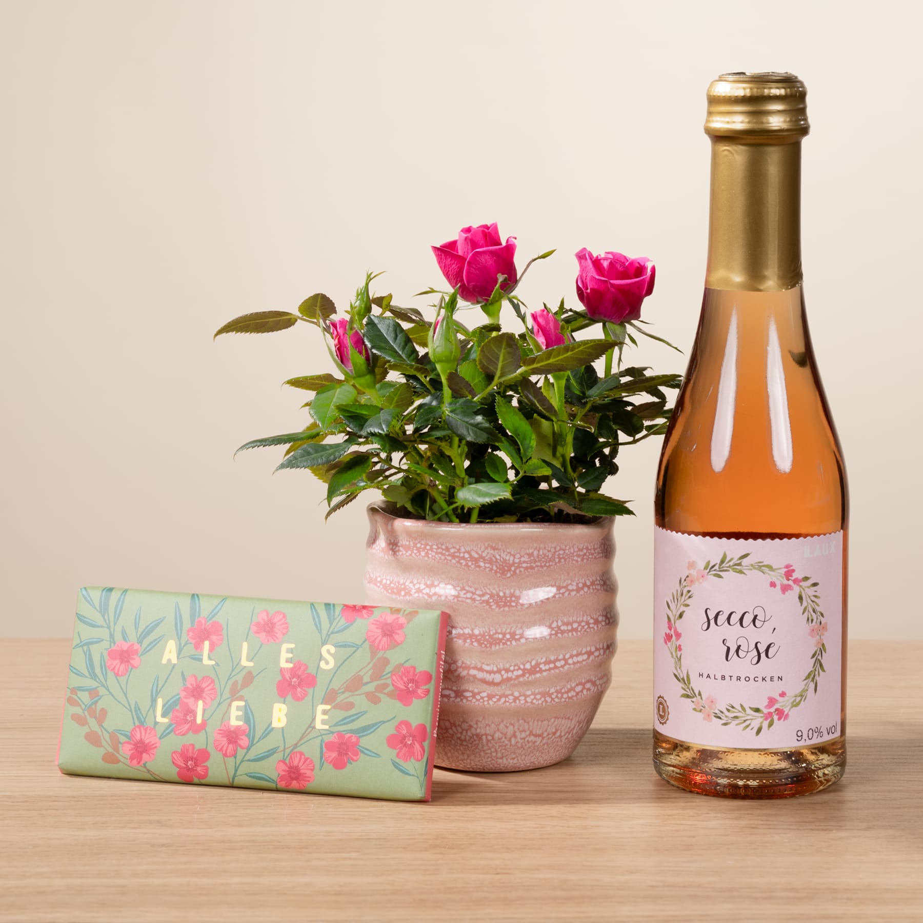 Ein Strauß leuchtend rosa Rosen in einem Keramiktopf steht auf einer Holzoberfläche, begleitet von einer Flasche Roséwein und einer dekorativen Geschenkbox mit Blumenmuster.