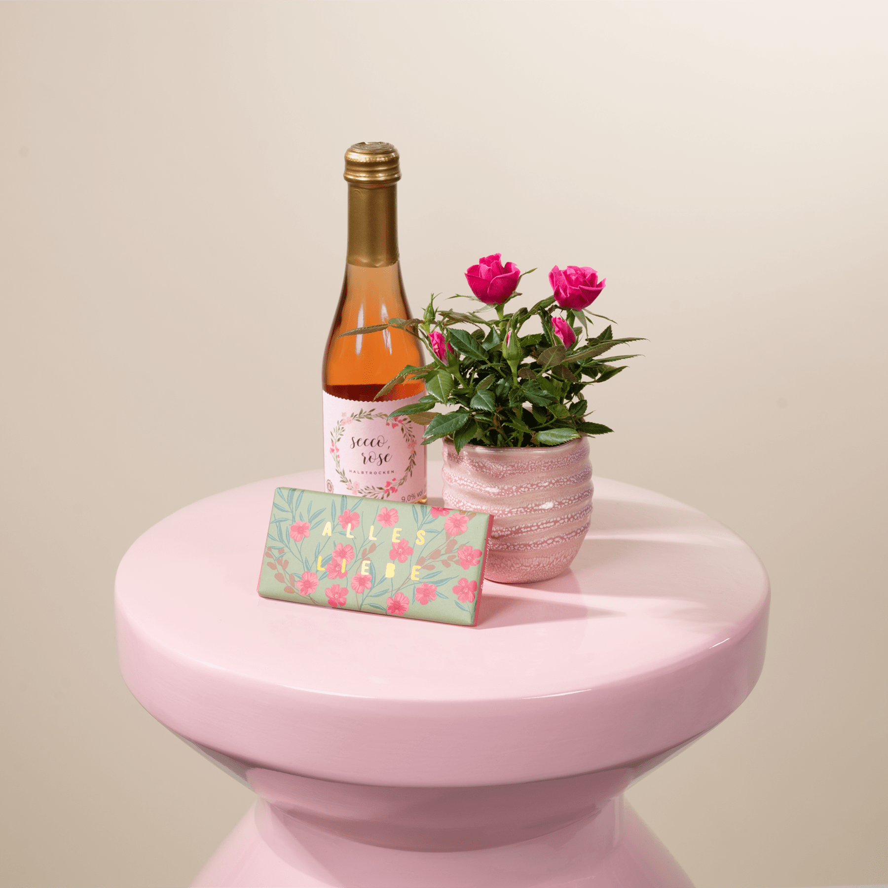 Auf einem rosafarbenen Sockel stehen eine Flasche mit orangefarbener Flüssigkeit, eine Topfpflanze mit rosafarbenen Blüten und eine Geschenkbox mit Blumenmuster.