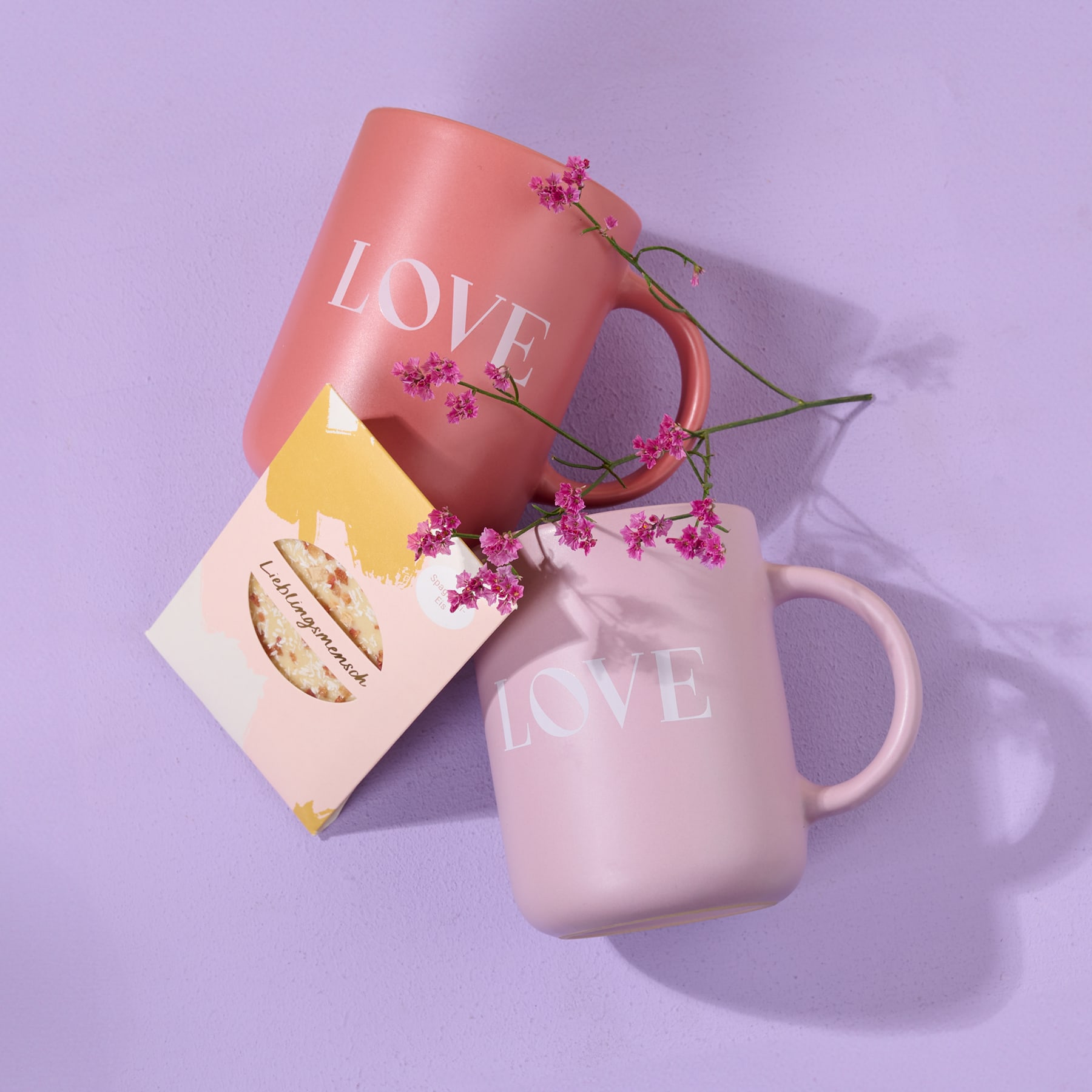 Vor einem zarten lavendelfarbenen Hintergrund stehen zwei Tassen mit dem Aufdruck „LOVE“ und einem kleinen Strauß rosa Blumen.