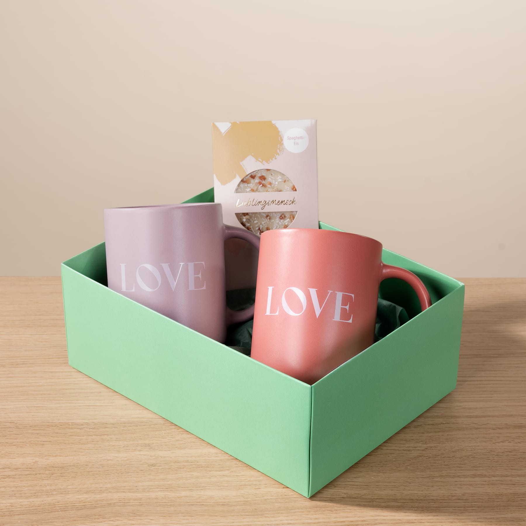 Eine grüne Schachtel mit zwei rosa Tassen mit dem Aufdruck „LOVE“ und einem kleinen Paket steht auf einer Holzoberfläche vor einem beigen Hintergrund.