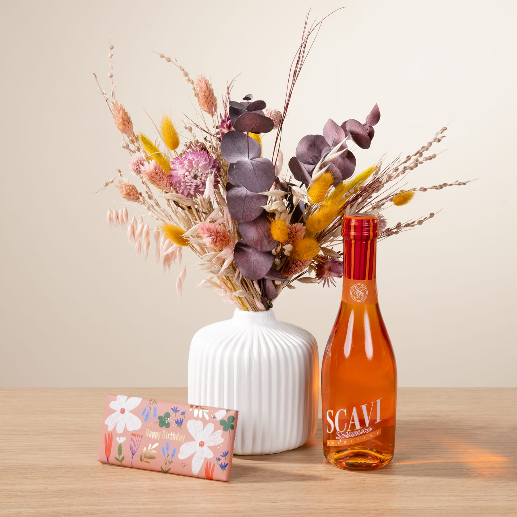 Ein farbenfroher Strauß getrockneter Blumen in einer weißen Vase, begleitet von einer Flasche orangefarbener Flüssigkeit und einer Geschenkbox mit Blumenmuster, alles vor einem neutralen Hintergrund.