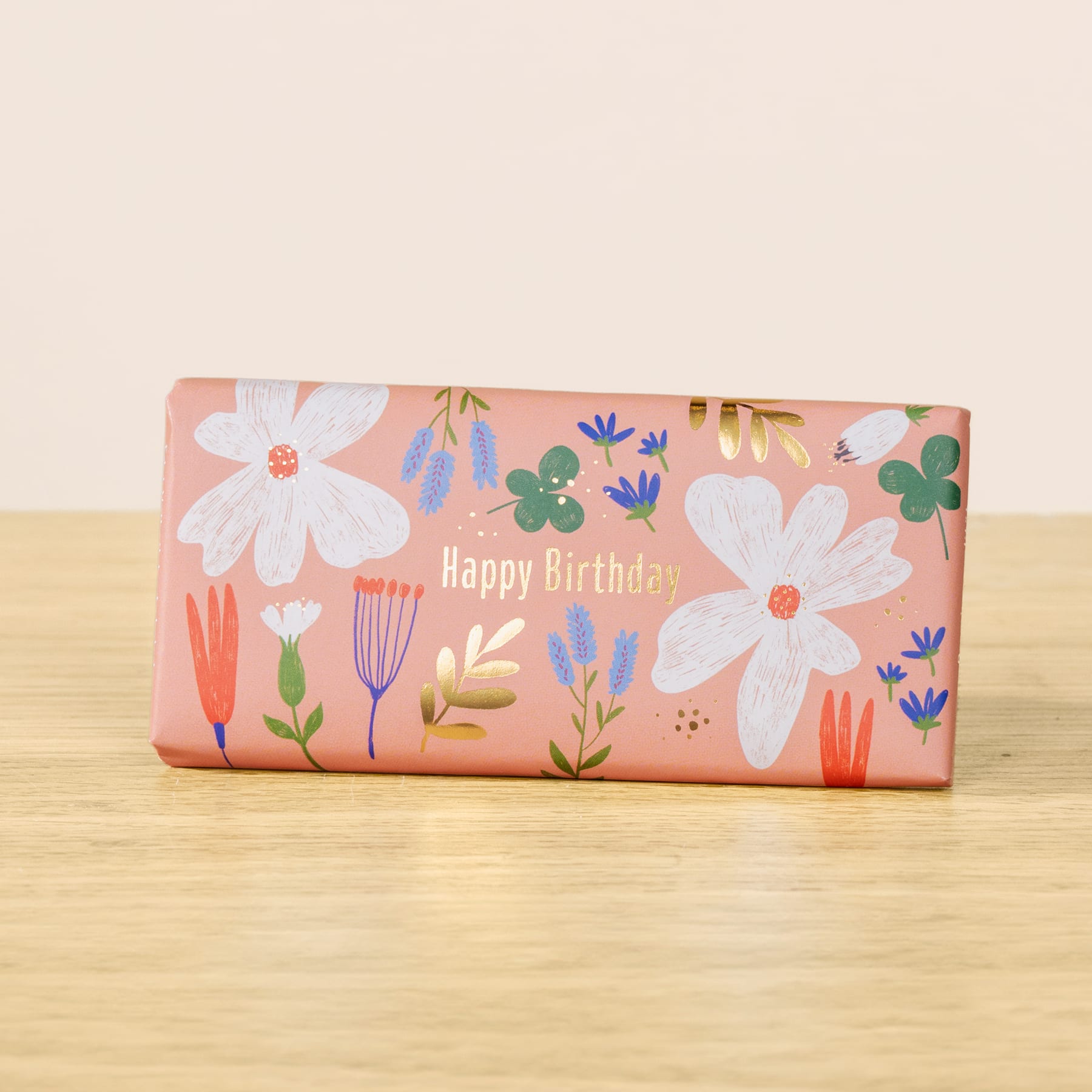 Eine rosa Geschenkbox mit Blumenmuster und dem Aufdruck „Happy Birthday“ steht auf einer Holzoberfläche.
