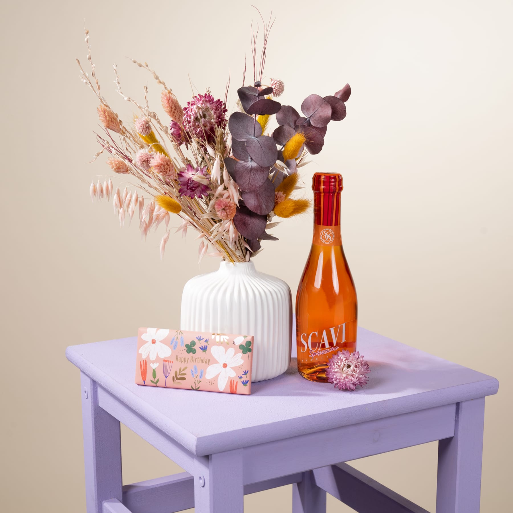 Auf einem lavendelfarbenen Beistelltisch steht eine Vase mit leuchtenden Trockenblumen, dazu eine Flasche Scavi-Wein und eine kleine dekorative Schachtel.