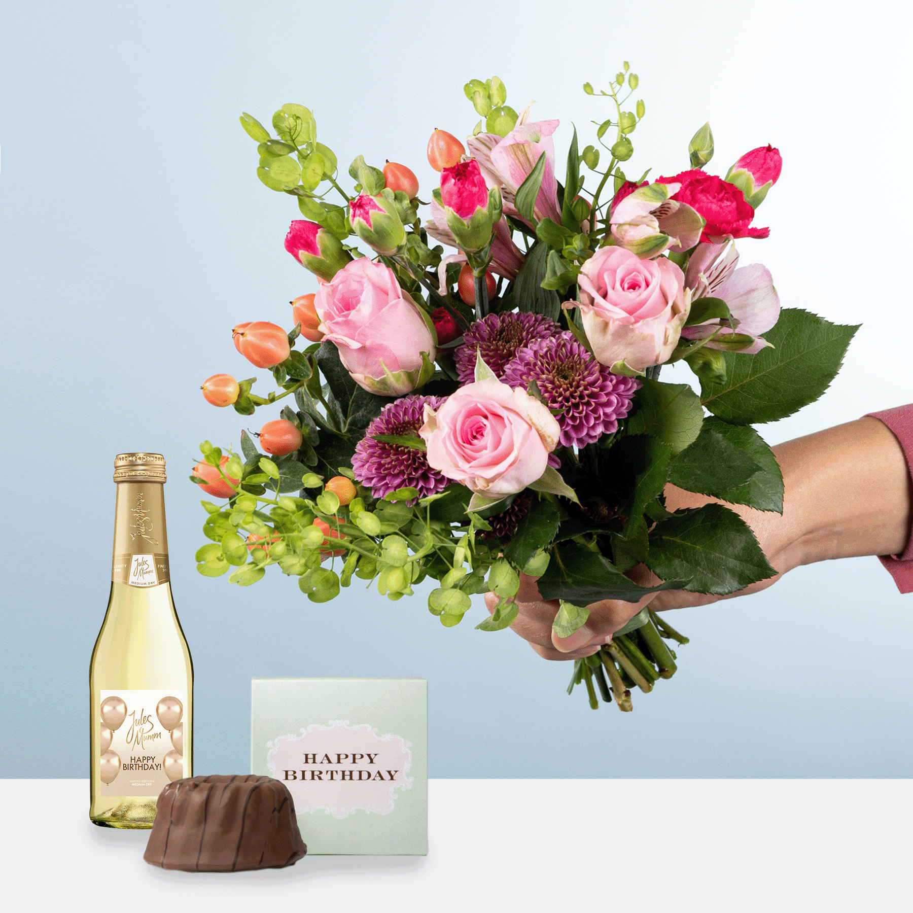 Eine Person hält einen farbenfrohen Strauß rosa Rosen, lila Chrysanthemen und Grünpflanzen in den Händen, begleitet von einer Flasche Champagner und einer Geburtstagskarte auf einem hellen Hintergrund.