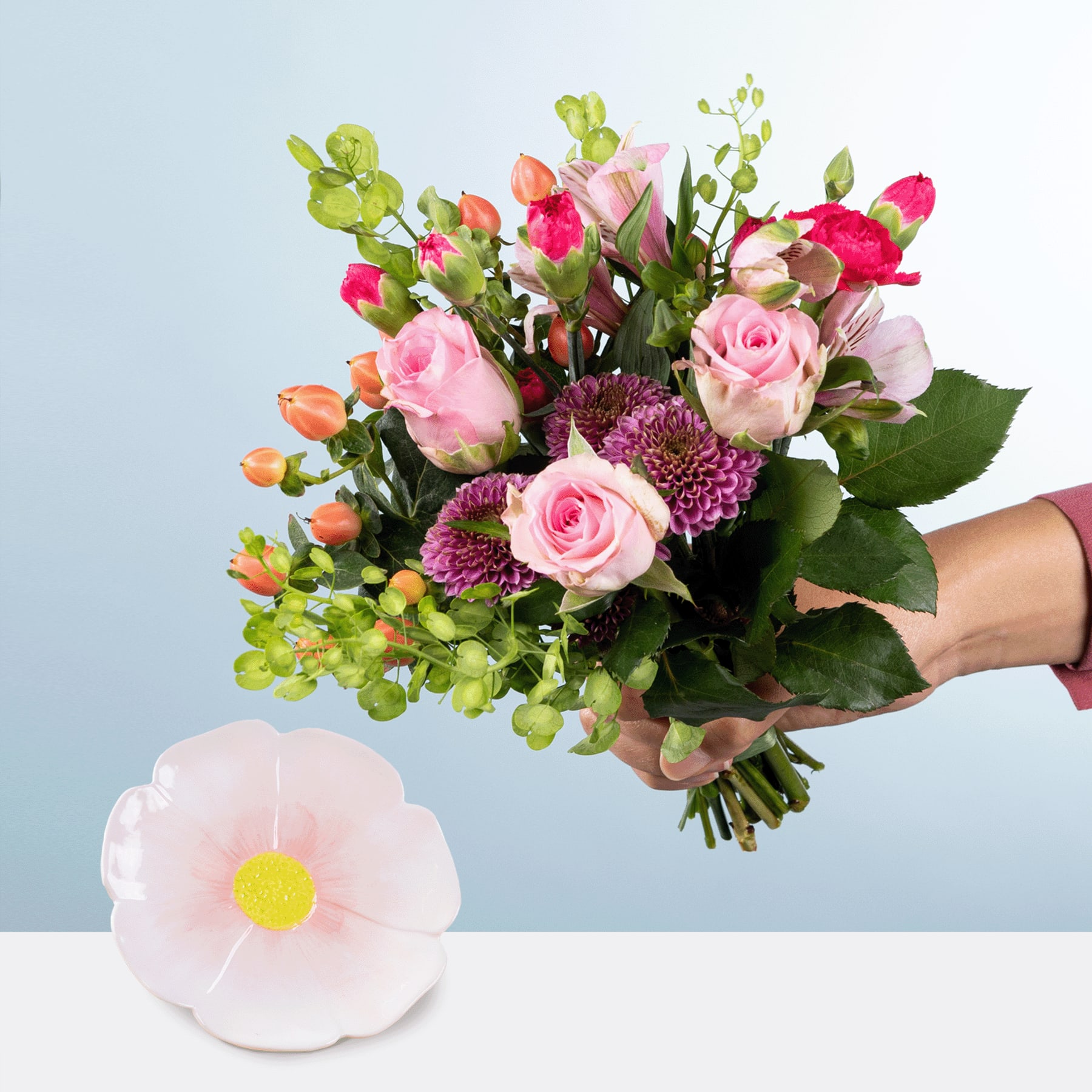 Ein leuchtender Strauß aus rosa Rosen, violetten Chrysanthemen und grünem Laub, den eine Person vor einem schlichten blauen Hintergrund in den Händen hält, mit einer einzelnen weißen Blume im Vordergrund.