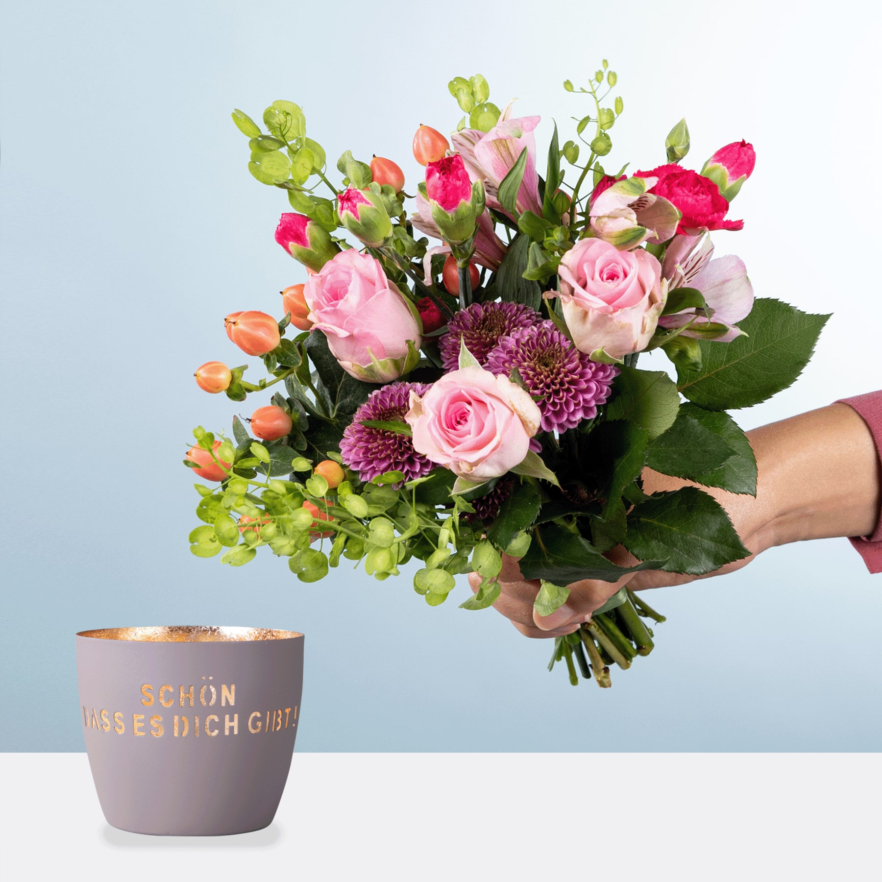 Ein leuchtender Strauß rosa Rosen, lila Chrysanthemen und grünem Laub in den Händen einer Person, mit einer grauen Keramiktasse mit deutschem Text im Vordergrund vor einem schlichten Hintergrund.