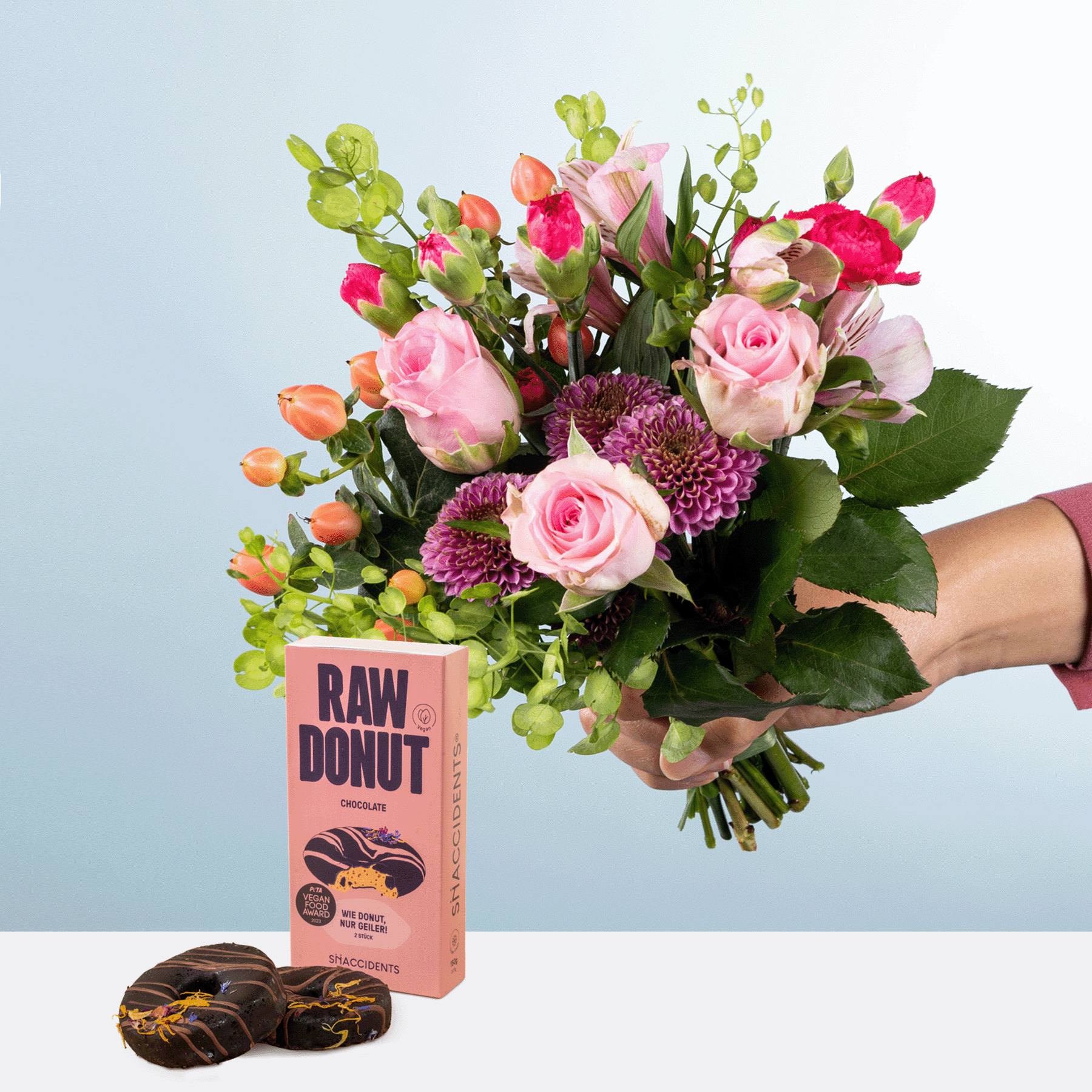Ein farbenfroher Strauß rosa Rosen, lila Chrysanthemen und anderer üppiger Grünpflanzen wird neben einer Schachtel roher Donuts auf einem hellblauen Hintergrund präsentiert.