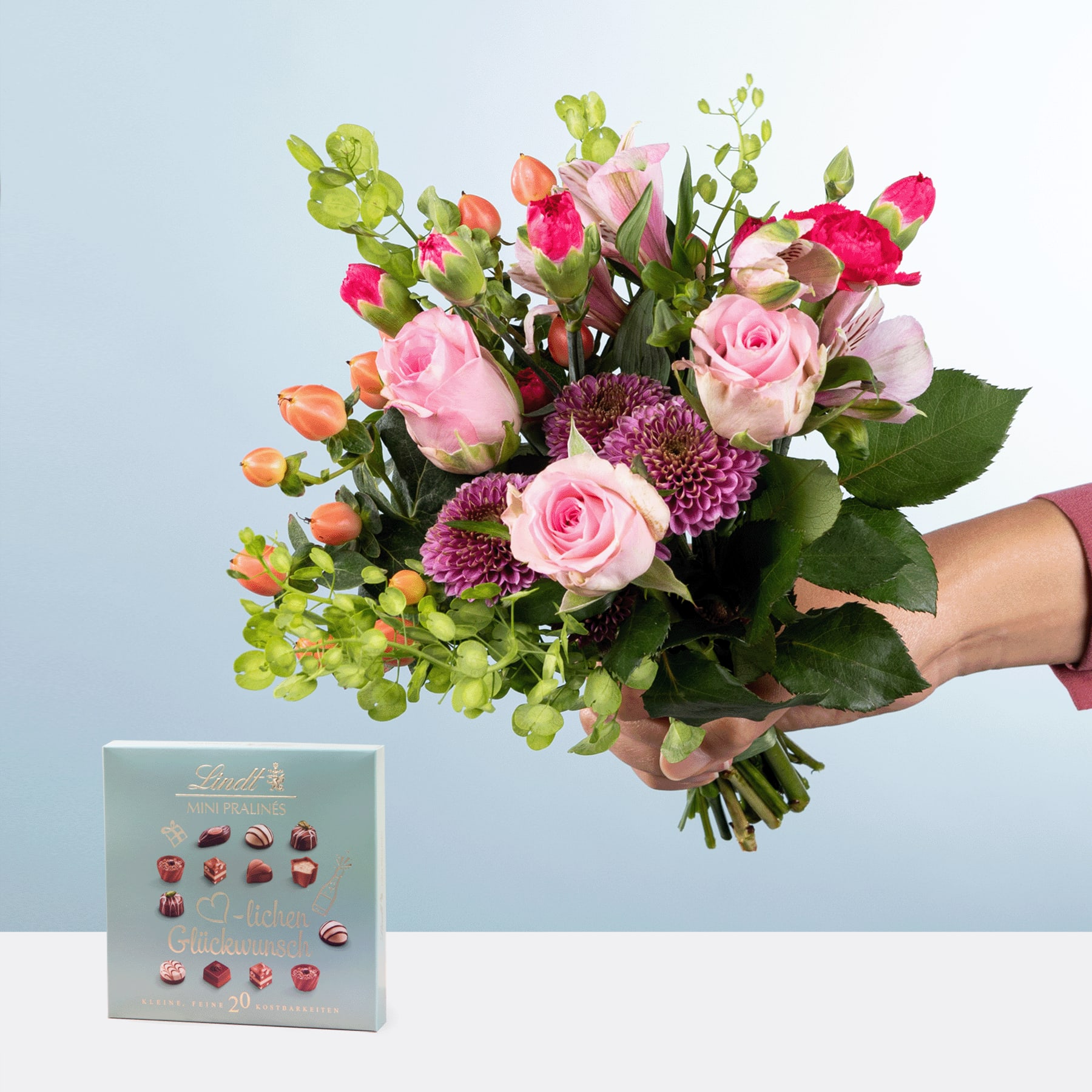 Eine Person hält einen farbenfrohen Strauß rosa Rosen, violetter Chrysanthemen und grünem Laub in den Händen. Der Strauß steht auf einem schlichten blauen Hintergrund, im Vordergrund ist eine kleine Geschenkbox zu sehen.