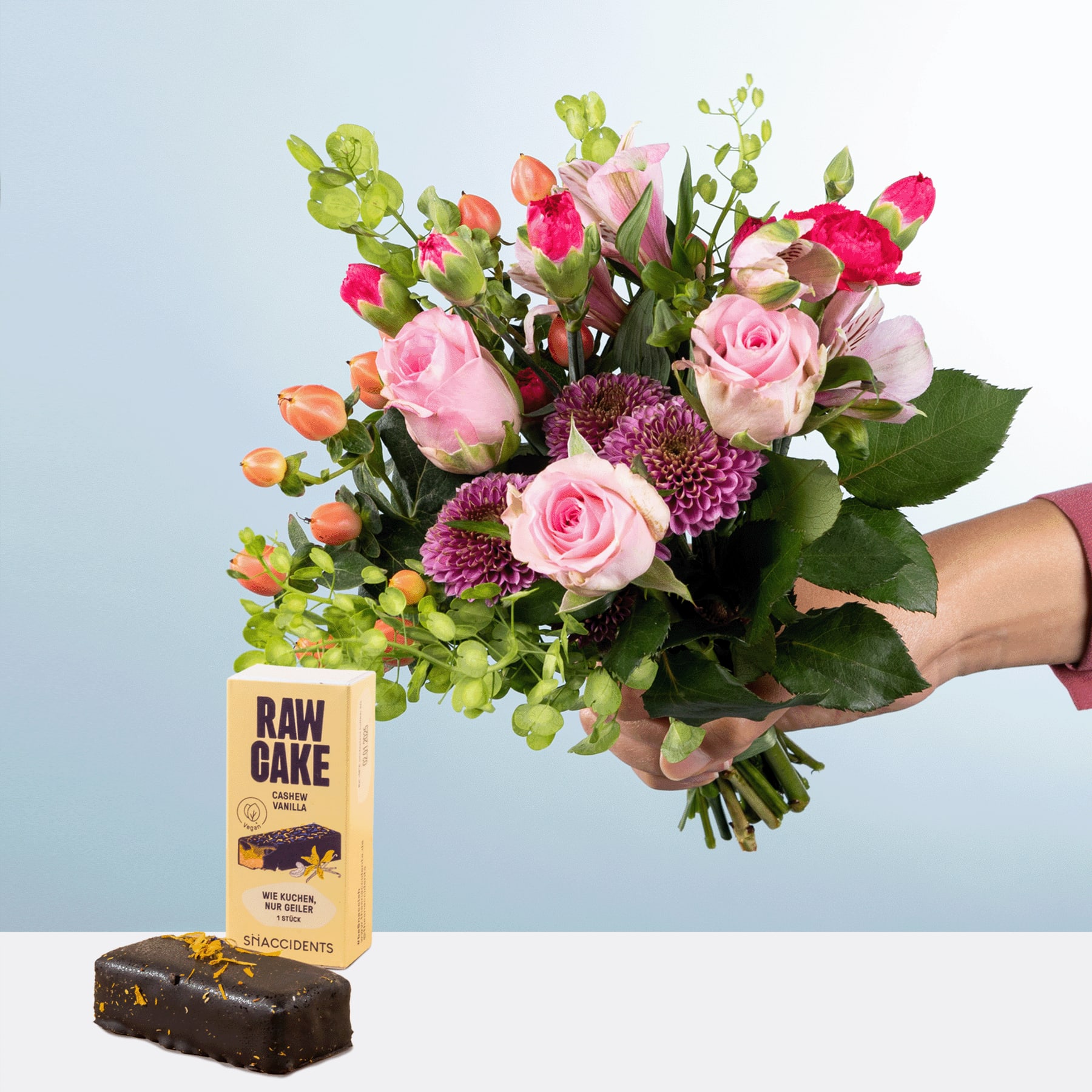 Ein farbenfroher Strauß aus rosa Rosen, violetten Chrysanthemen und Grünpflanzen wird vor einer Schachtel mit Rohkuchen gehalten, vor einem schlichten Hintergrund.