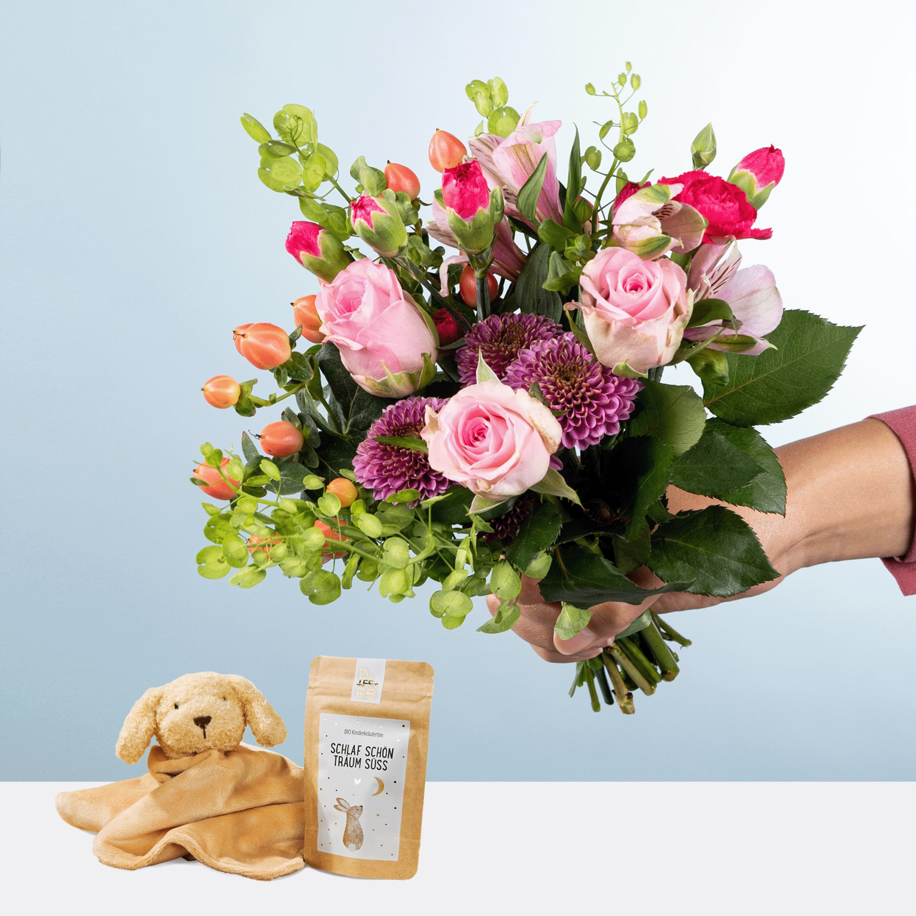 Eine Person hält einen farbenfrohen Strauß rosa Rosen, lila Chrysanthemen und Grünpflanzen in den Händen, im Hintergrund sind ein Plüschteddybär und eine kleine Geschenkbox zu sehen.