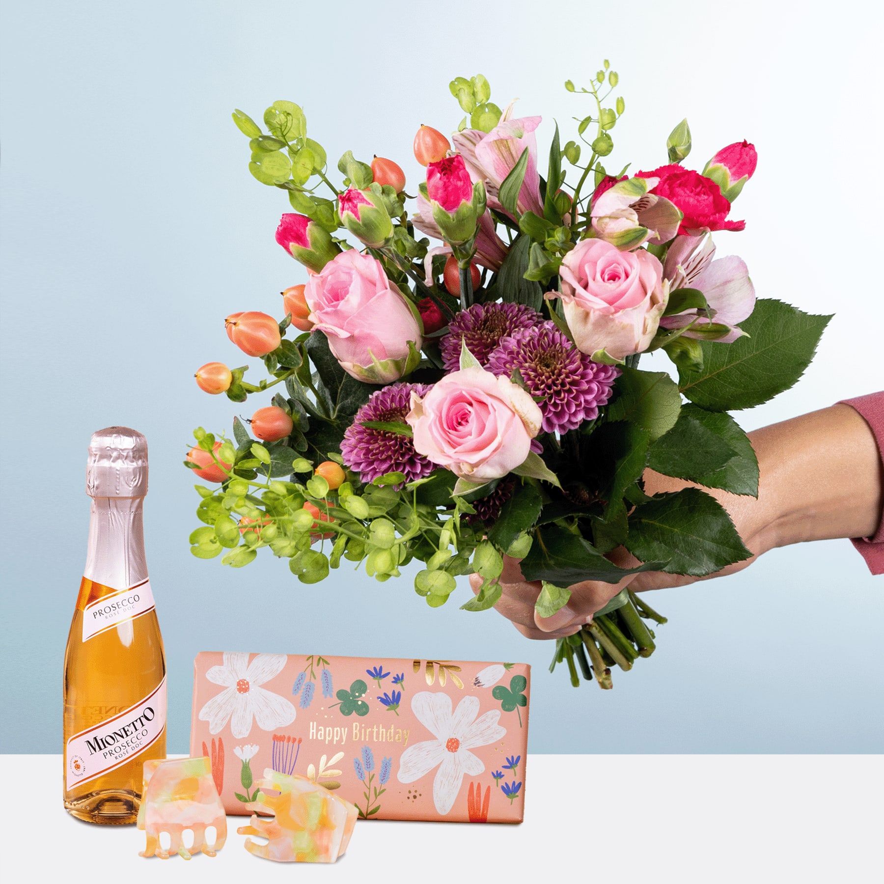 Ein leuchtender Strauß aus rosa Rosen, lila Chrysanthemen und Grünpflanzen, gehalten von einer Person in der Hand, begleitet von einer Flasche Champagner und einer Geschenkbox mit Blumenmuster auf hellblauem Hintergrund.