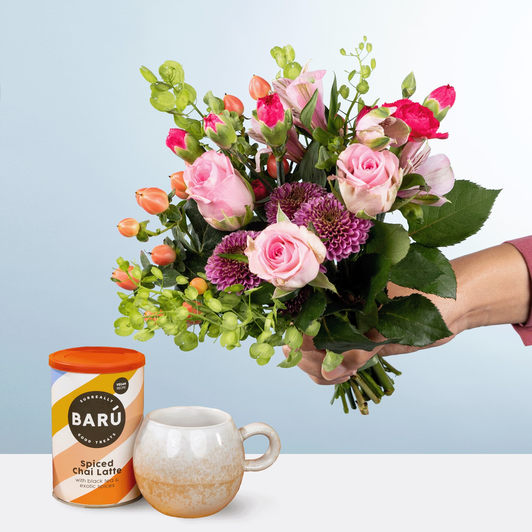Eine Person hält einen farbenfrohen Strauß rosa Rosen, violetter Chrysanthemen und grünem Laub in den Händen, im Hintergrund steht auf dem Tisch eine Dose Baru-Kokosmilch.