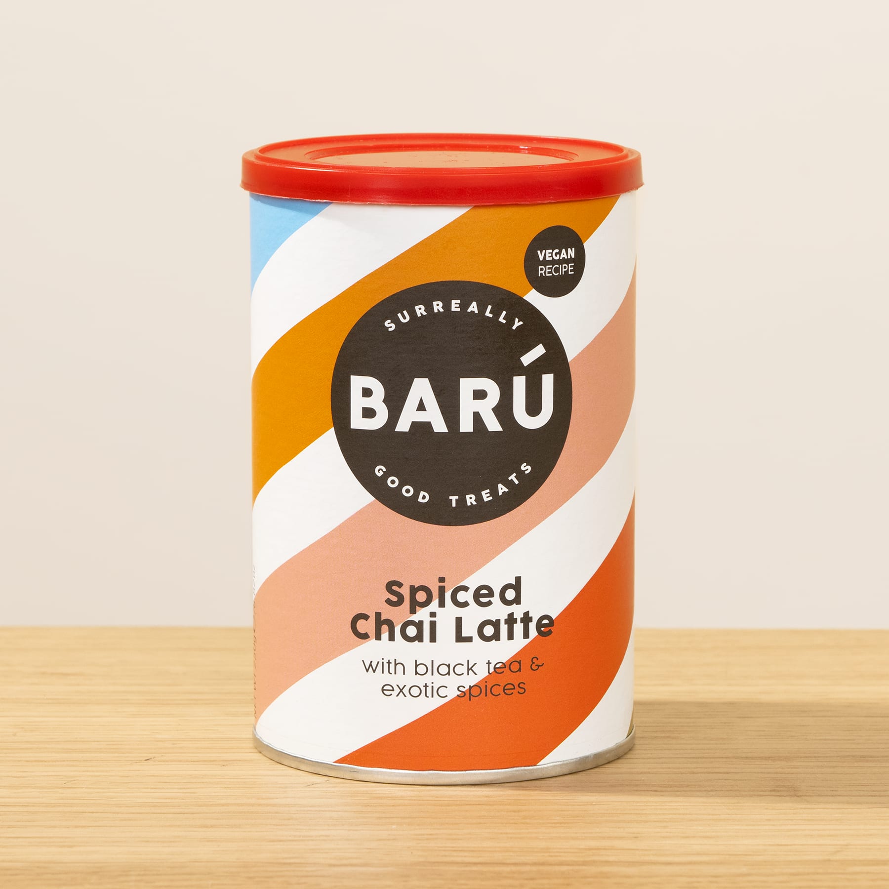 Eine bunte Blechdose mit der Aufschrift „Baru Spiced Chai Latte“ steht auf einer Holzoberfläche vor einem schlichten Hintergrund.