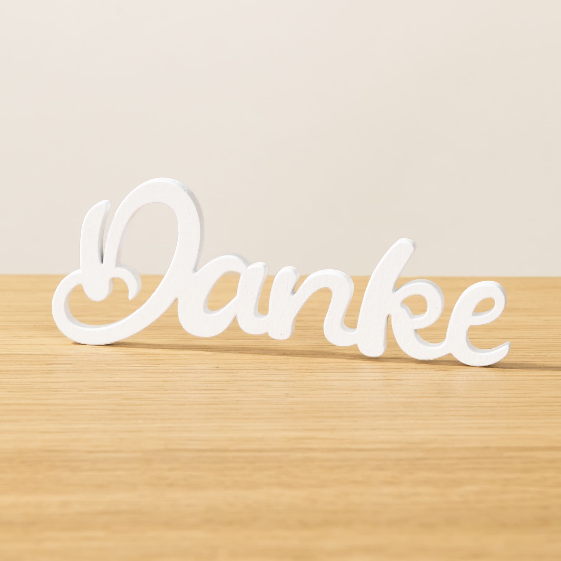 Das Bild zeigt das Wort „Danke“ in weißer Schreibschrift auf einer Holzoberfläche vor hellem Hintergrund.