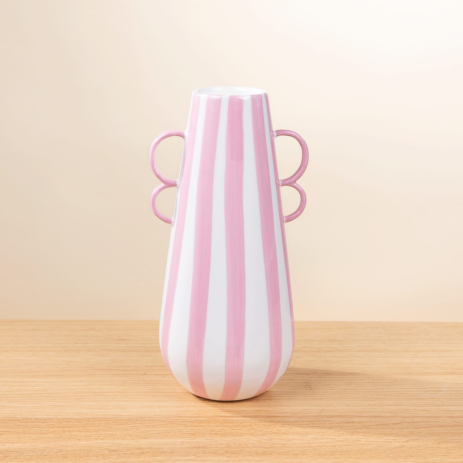 Eine rosa-weiß gestreifte Keramikvase mit schmetterlingsförmigen Griffen steht auf einem Holztisch vor einem schlichten beigen Hintergrund.