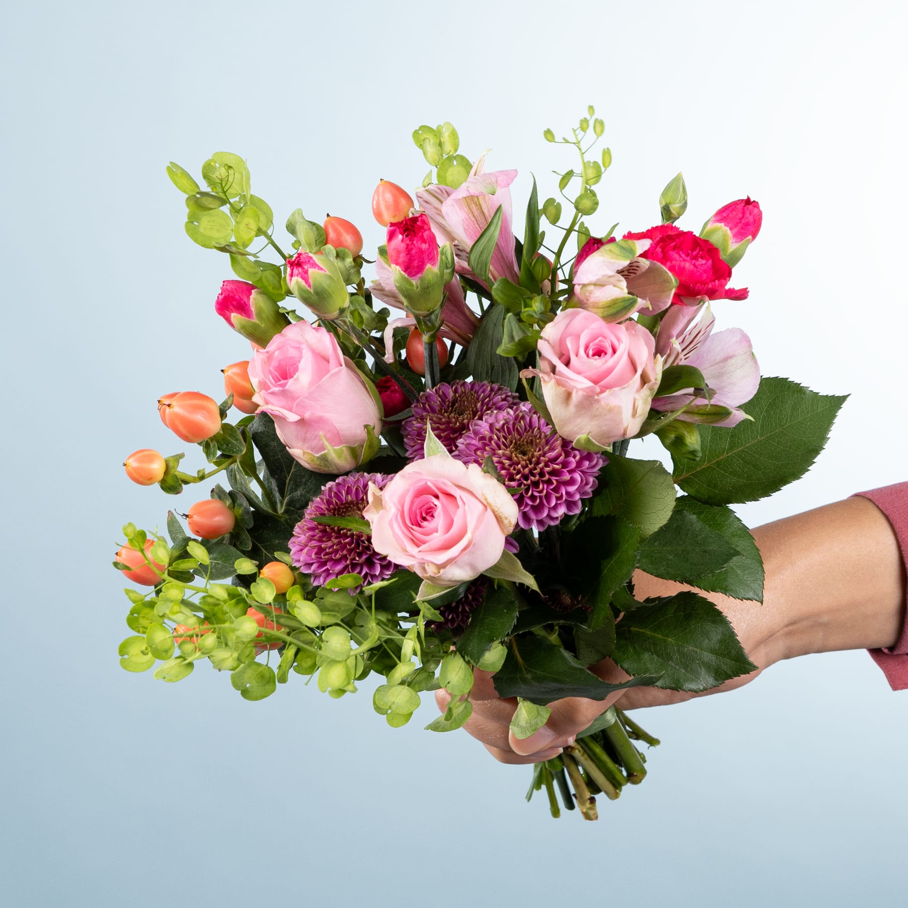 Ein leuchtender Strauß aus rosa Rosen, grünem Laub und anderen bunten Blumen, den eine Person vor einem schlichten hellblauen Hintergrund in den Händen hält.