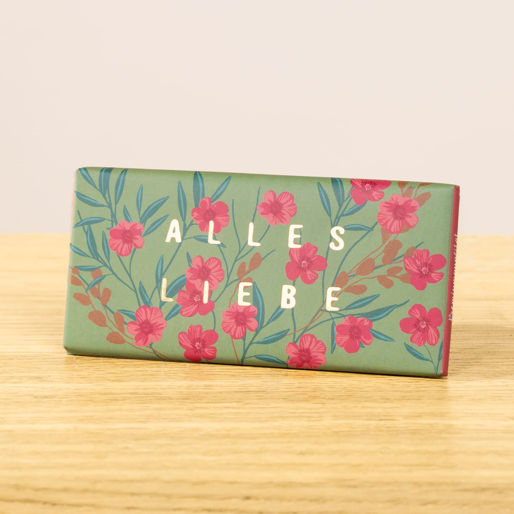 Eine grüne rechteckige Schachtel mit dem Text „ALLES LIERE“ und einem floralen Muster aus roten Blumen und grünen Blättern auf hellem Hintergrund.