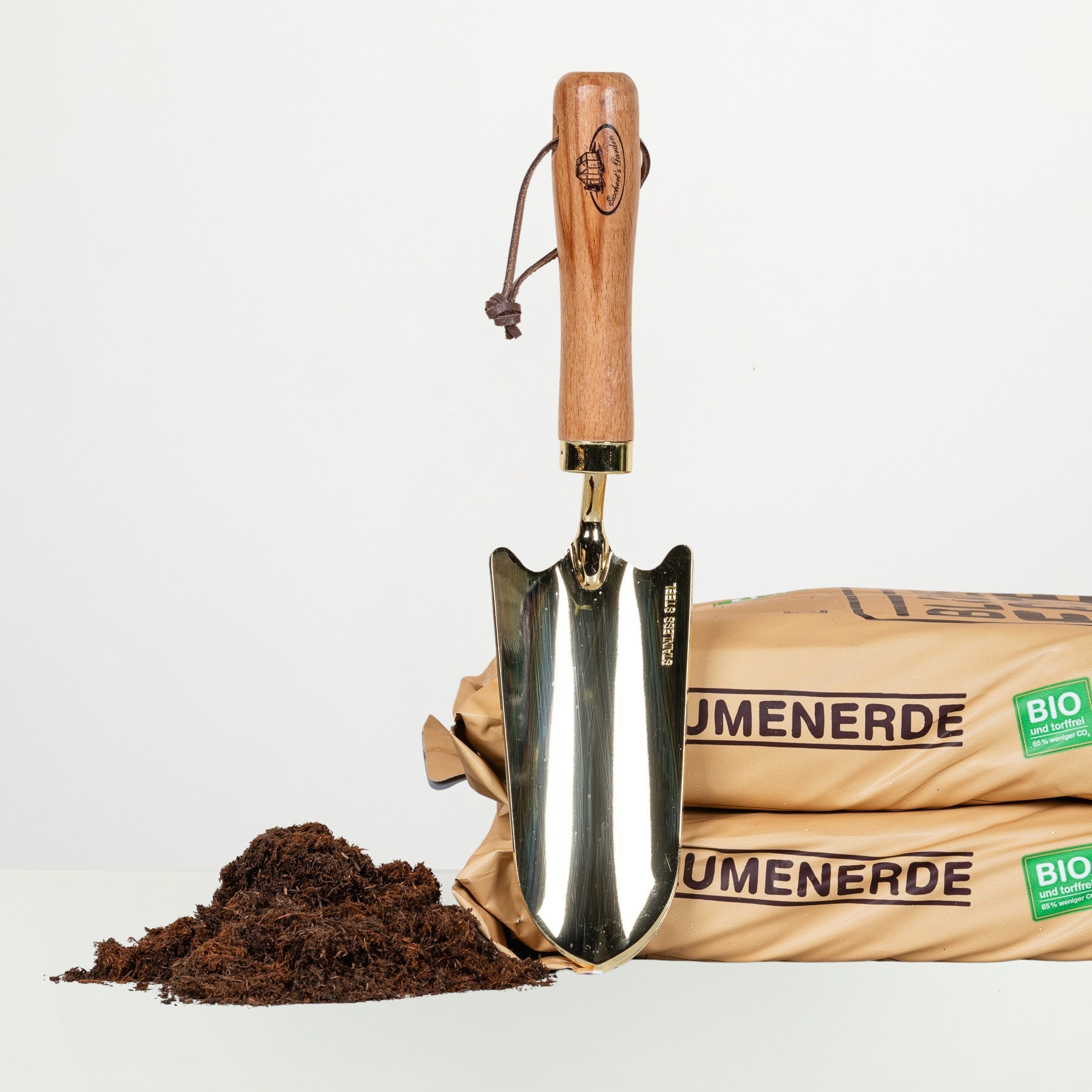 Ein Gartengerät mit Holzgriff, wahrscheinlich eine Kelle oder eine kleine Schaufel, liegt neben zwei Säcken mit Bioerde oder Kompost mit der Aufschrift „Jmenerde Bio“.
