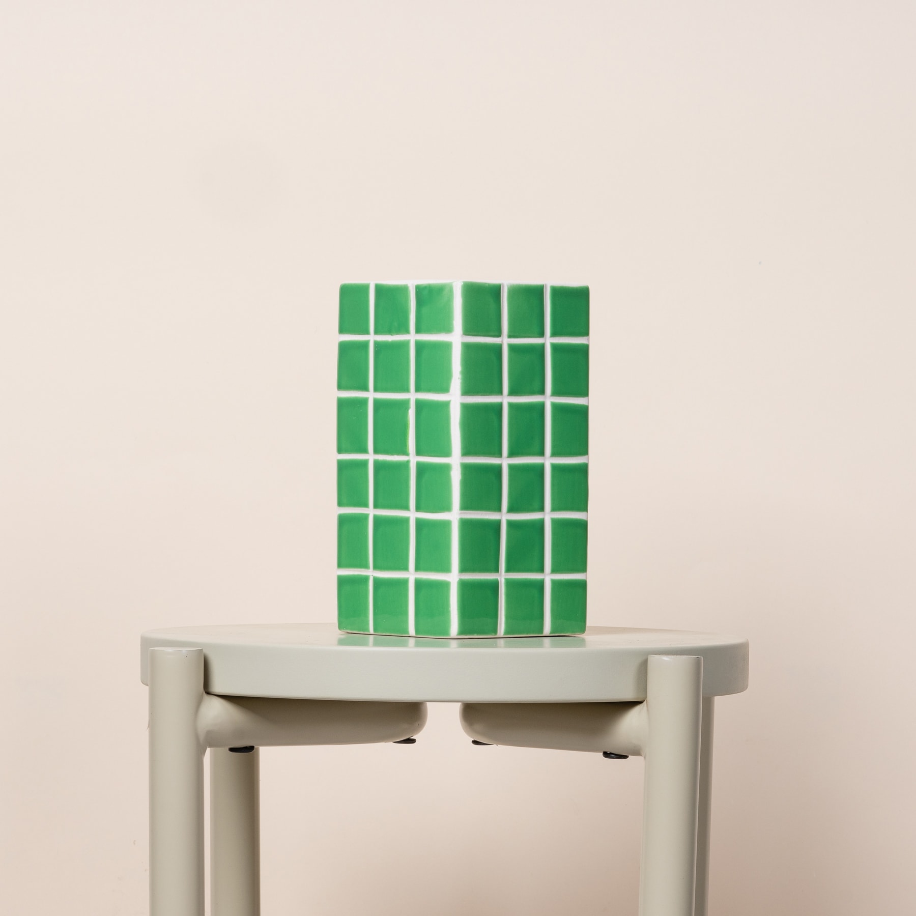 Eine grüne mosaikartige Struktur steht auf einem weißen Sockel vor einem schlichten beigen Hintergrund.