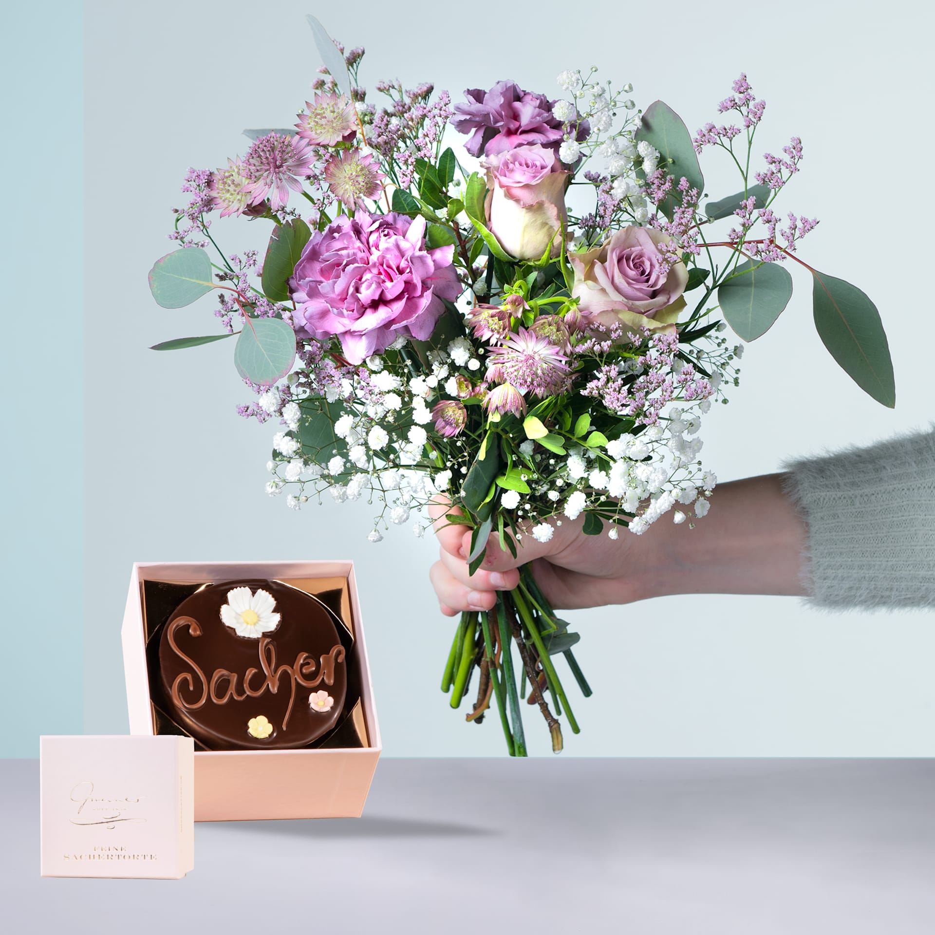 Eine Person hält einen farbenfrohen Strauß rosa und weißer Blumen, darunter Rosen und Pfingstrosen, in den Händen, zusammen mit einer kleinen Pralinenschachtel mit der Aufschrift „Sasher“.