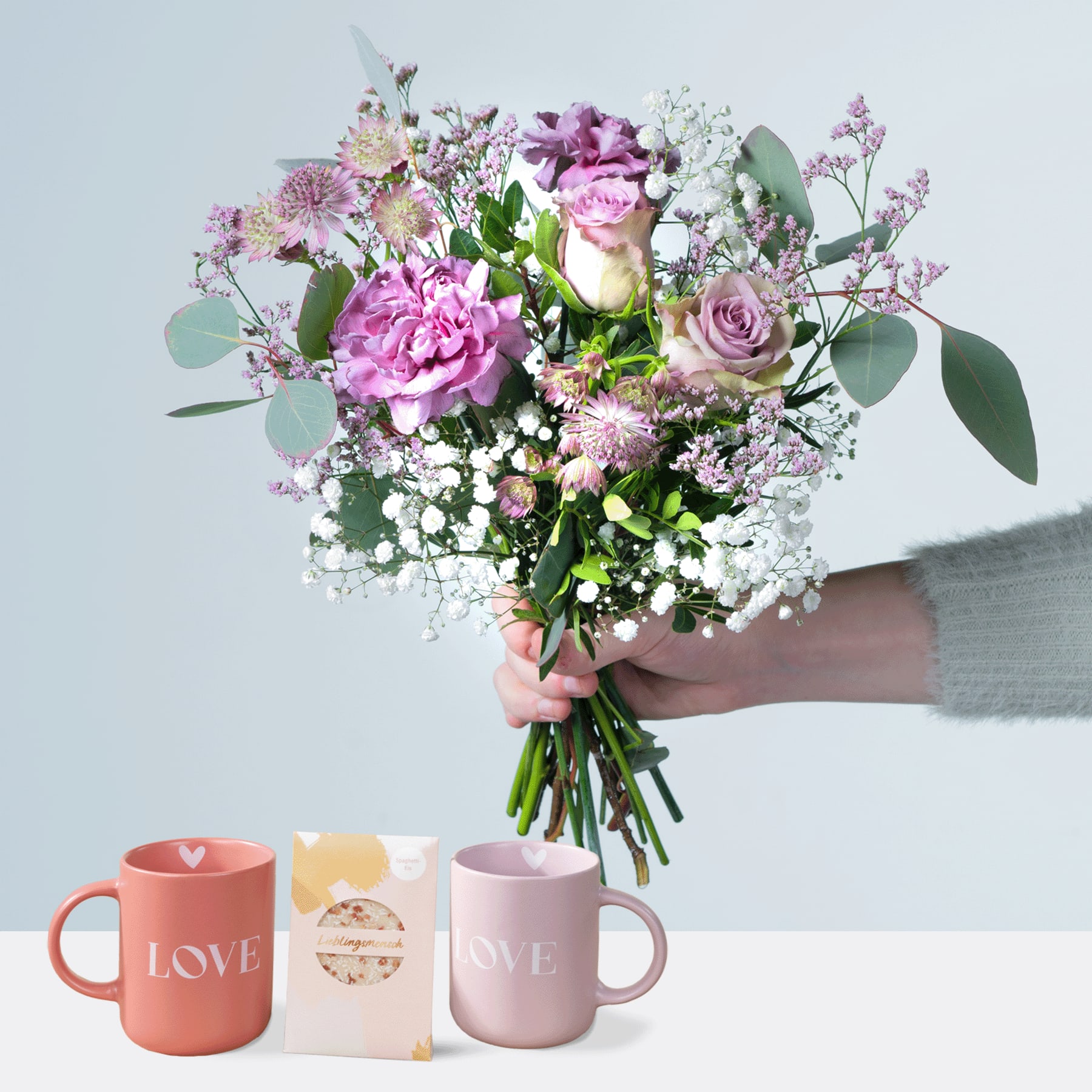 Eine Person hält einen farbenfrohen Strauß rosa und violetter Blumen, darunter Rosen und Pfingstrosen, in den Händen, begleitet von zwei Tassen mit der Aufschrift „LOVE“ vor einem schlichten Hintergrund.