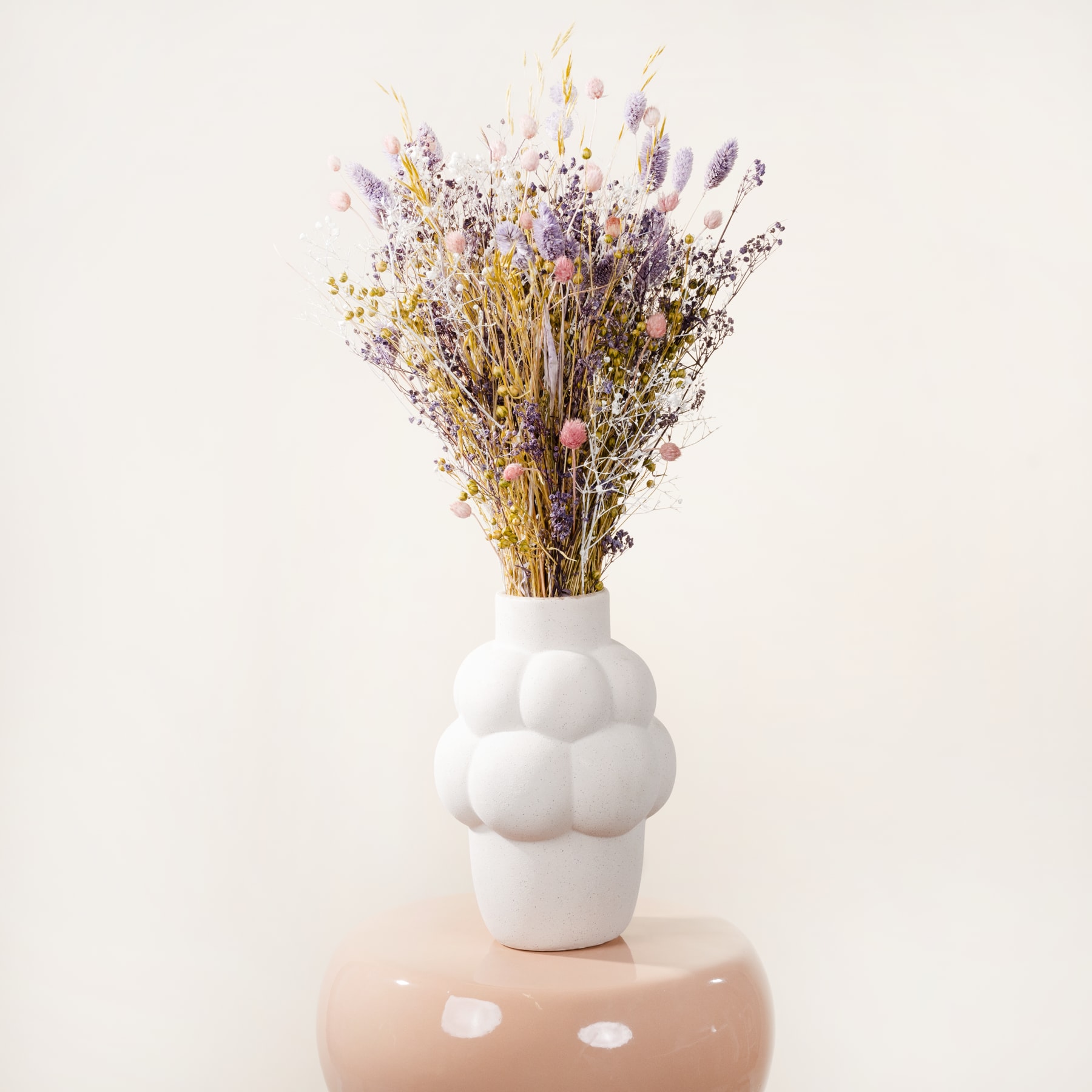 Ein Strauß leuchtender Trockenblumen in einer weißen Keramikvase steht auf einer blassrosa Oberfläche mit weißen Punkten.