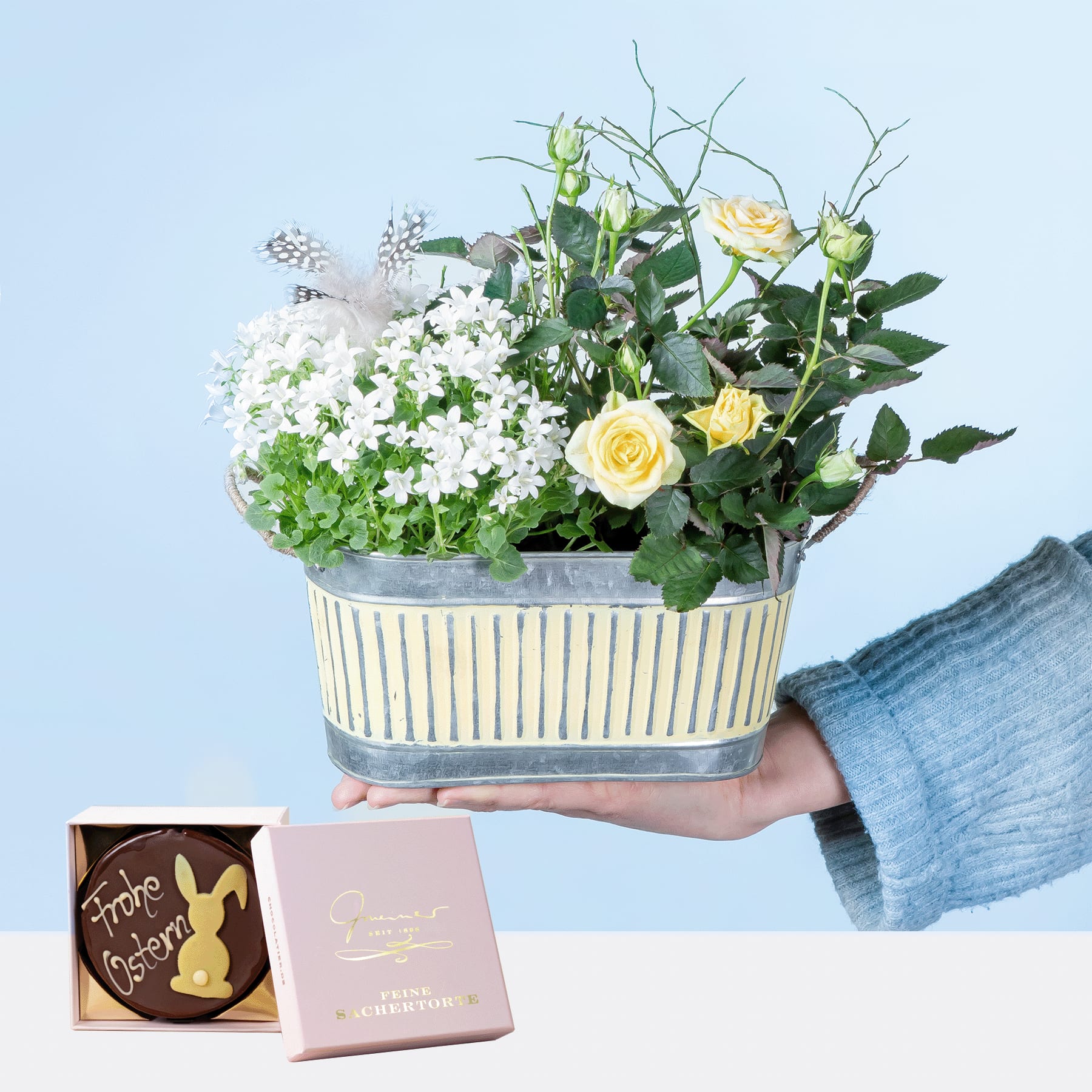 Eine Person hält einen Strauß weißer und gelber Blumen in einem gestreiften Behälter in den Händen. Im Vordergrund ist auf hellblauem Hintergrund eine Schachtel Pralinen zu sehen.